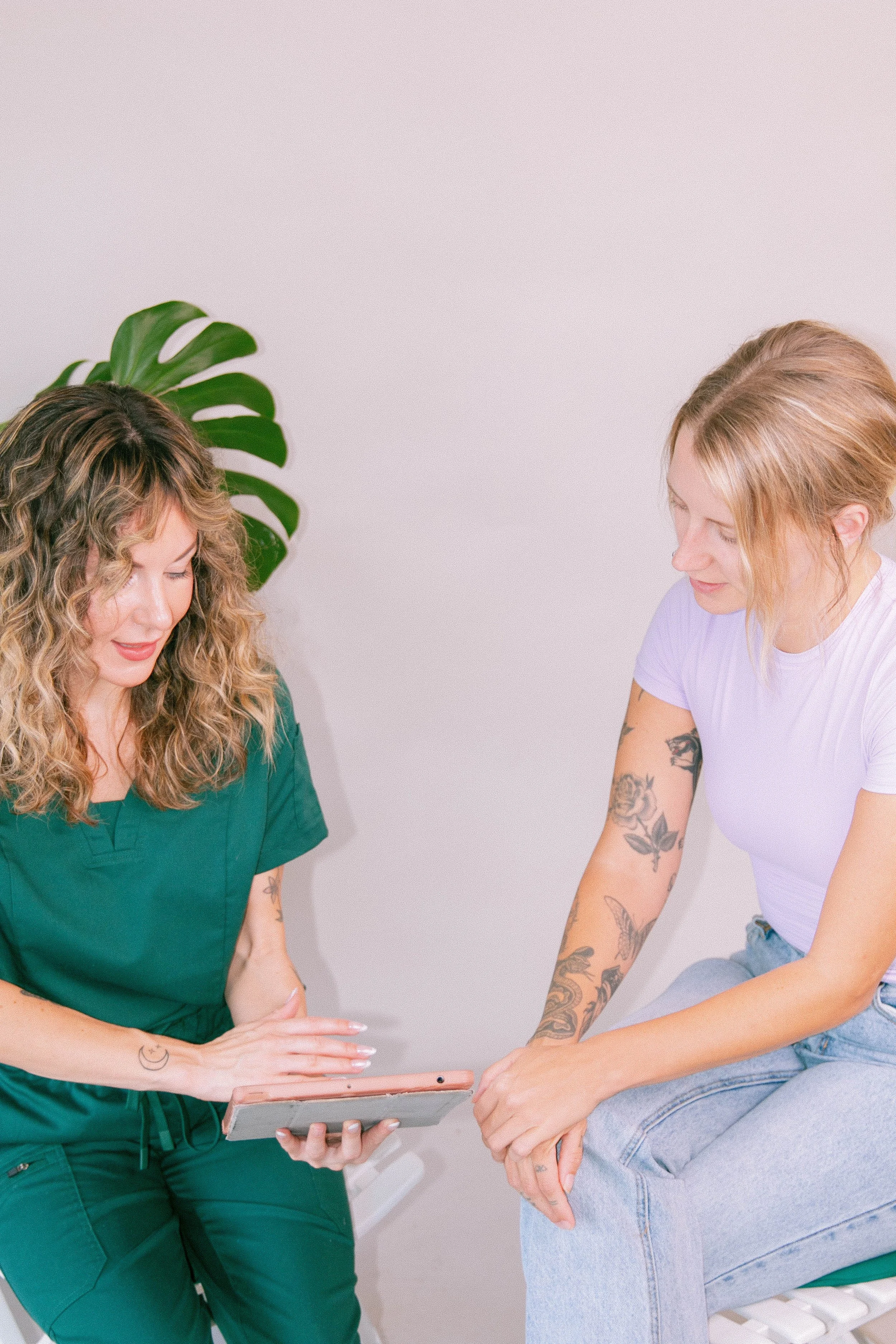 Two women sitting together, looking at a tablet, with one woman in green scrubs and the other in a white t-shirt with tattoos on her arms, in a room with a large green monstera plant in the background.