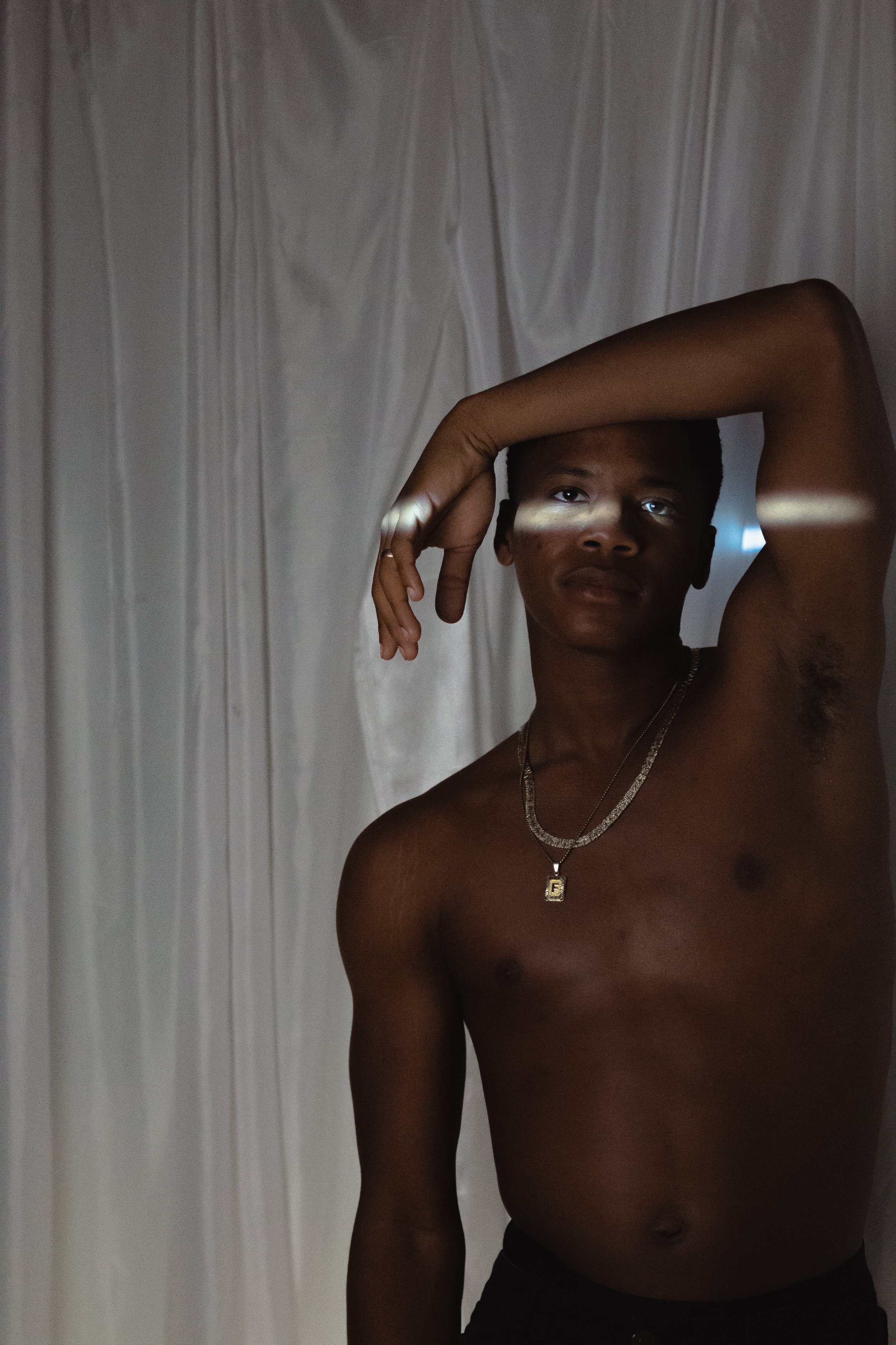 A shirtless young man with a dark complexion poses with his right arm bent over his head. He has short hair and is wearing a chain necklace with a pendant and a ring on his finger. The background is a plain, light-colored curtain.