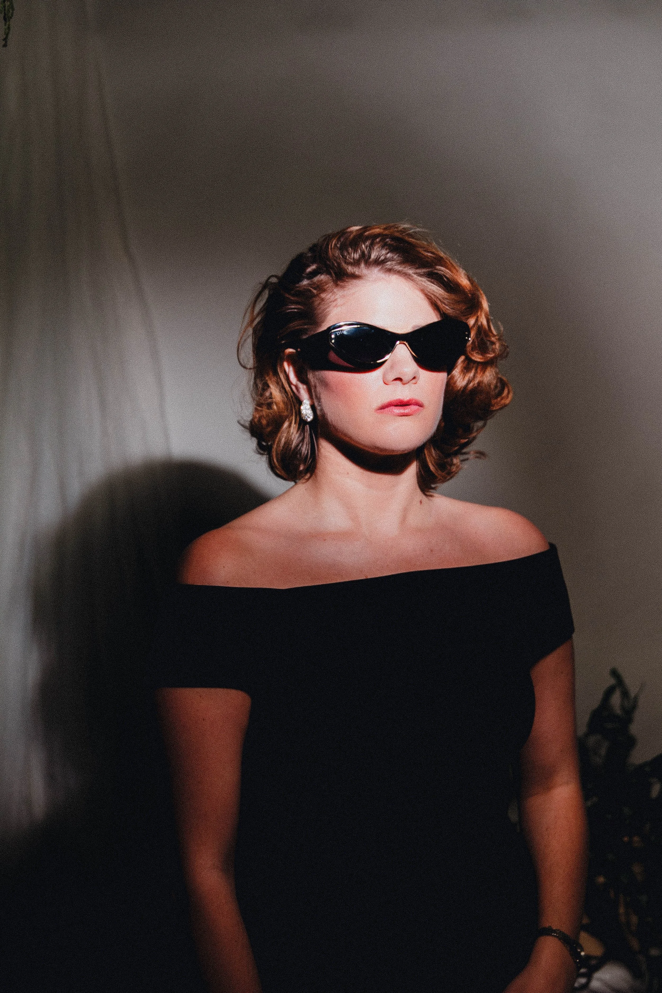 Woman with short curly brown hair wearing large black sunglasses, diamond earrings, black off-shoulder top, and a bracelet on her left wrist, standing indoors against a plain wall.