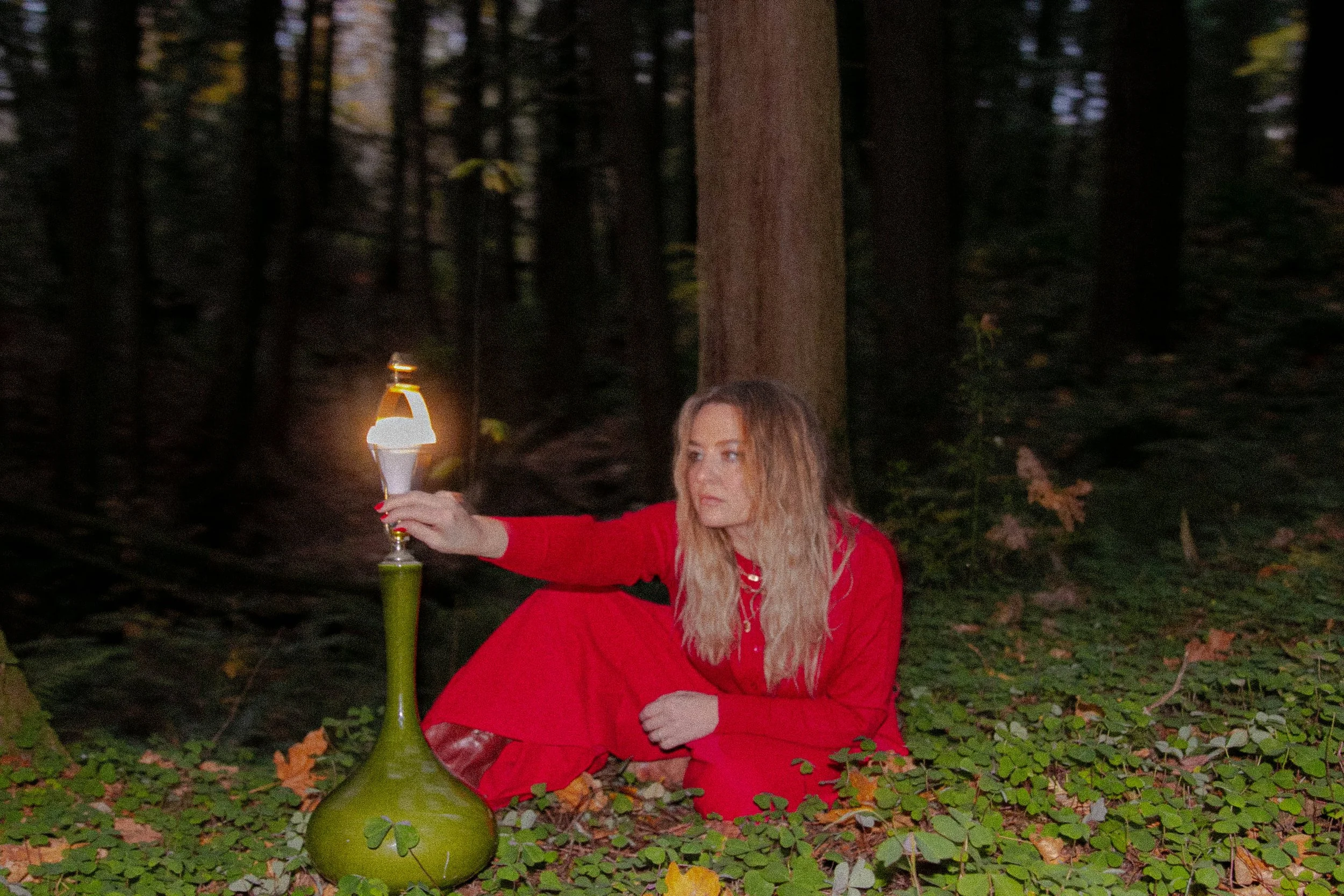 A woman with long blonde hair wearing a red coat, sitting on the ground in a forest at night, holding a lit lantern, with a large green bottle on the ground in front of her.