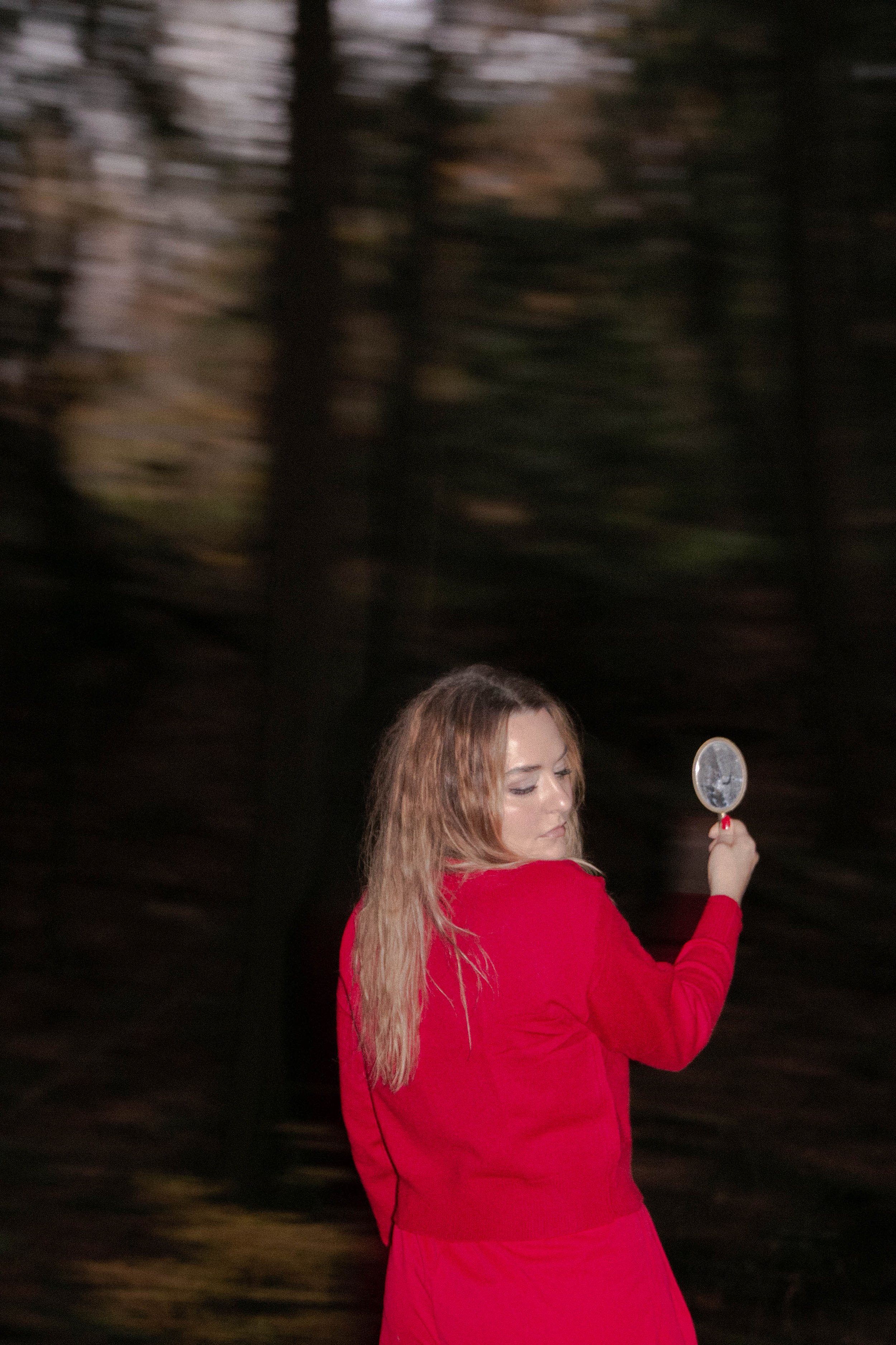 A woman with long, light brown hair wearing a red dress holds a circular mirror with a decorative frame, standing against a dark, blurred background.