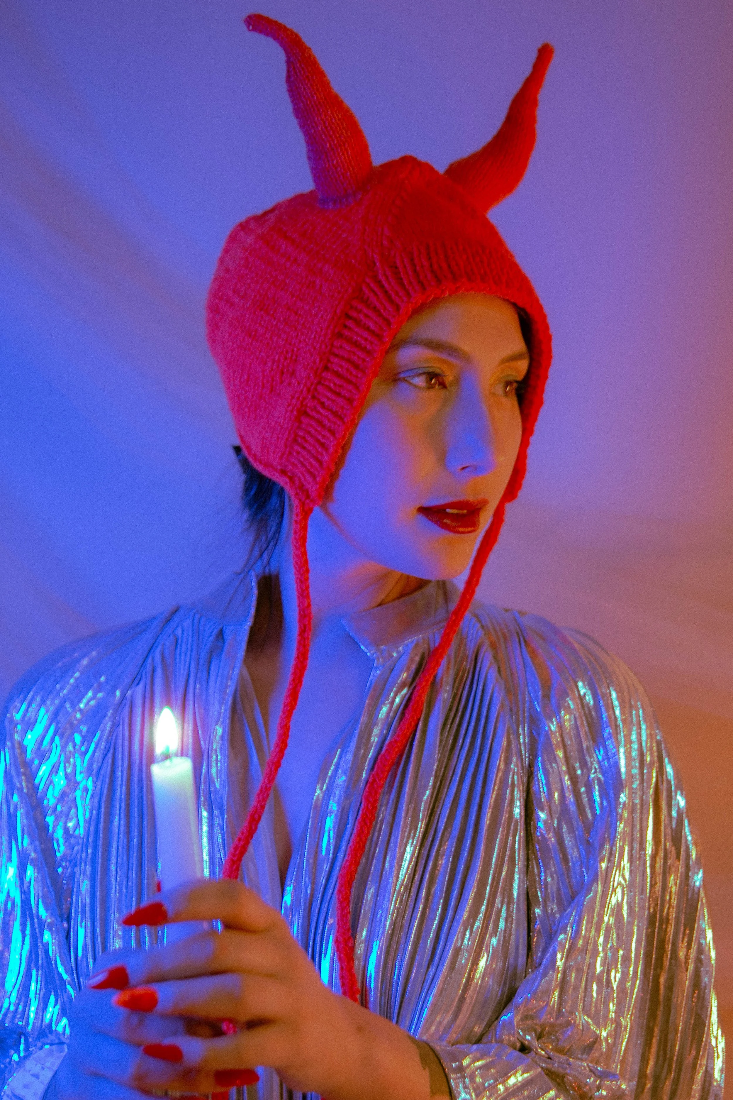 A woman wearing a red knit hat with two tall, pointed horns. She is holding a lit candle with a matchstick. She is wearing a shiny, iridescent jacket, and the background has colorful, soft lighting.