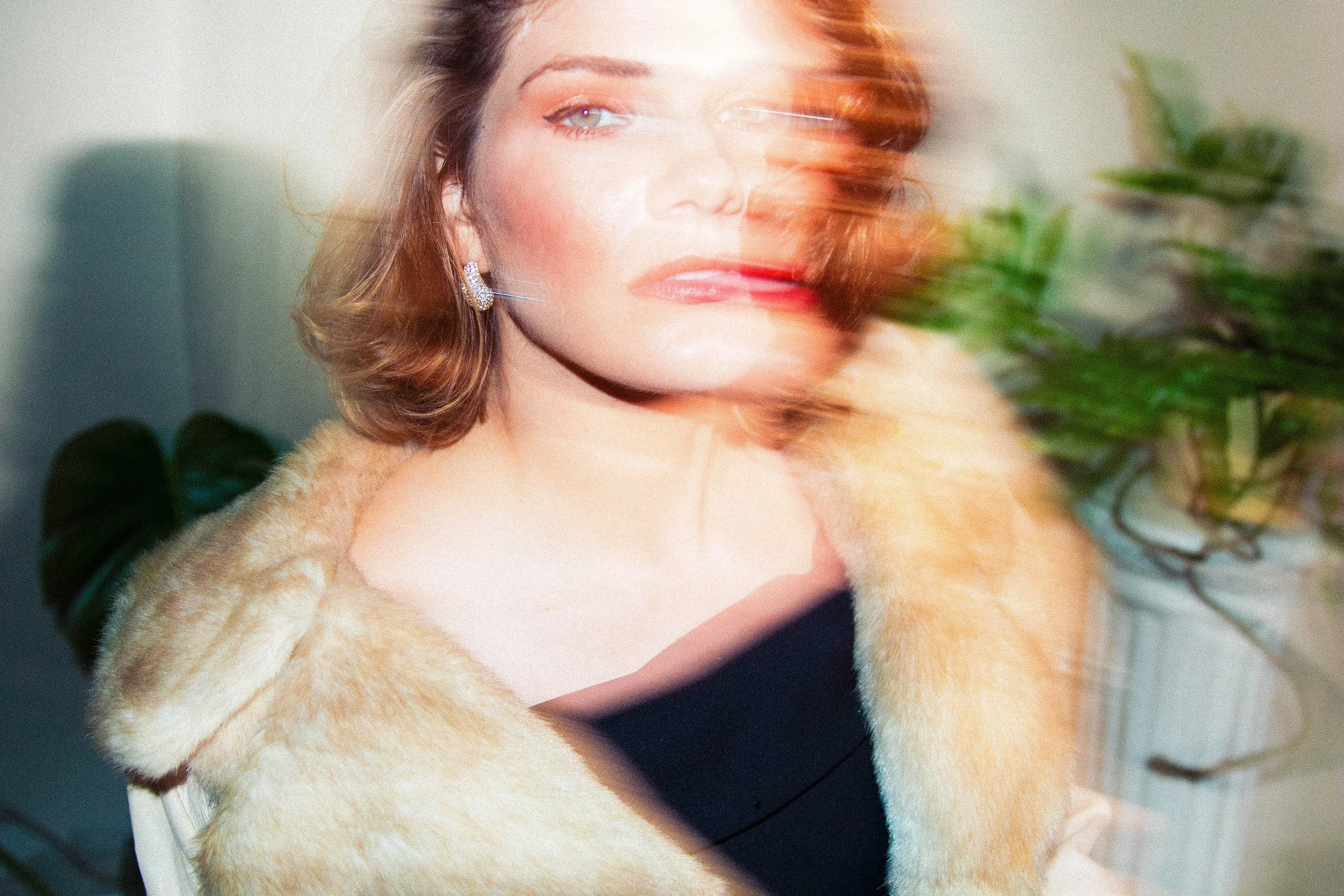 A woman with light skin and shoulder-length, wavy hair, dressed in a beige fur coat and black top, looking off to the side in a well-lit setting with green plants in the background.