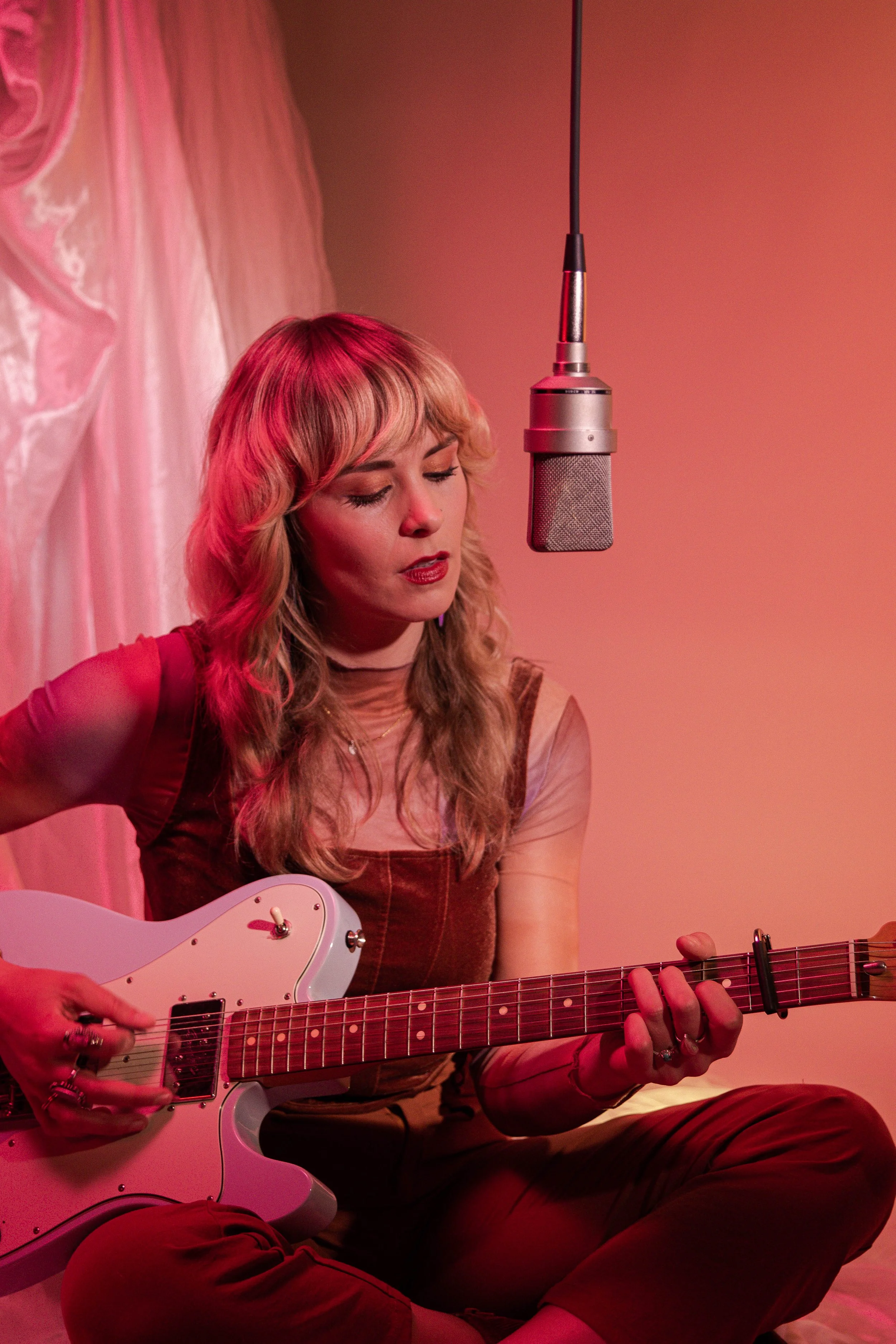 Young woman with wavy blonde hair playing an electric guitar and singing into a microphone in a cozy, softly lit room with pink and red tones.