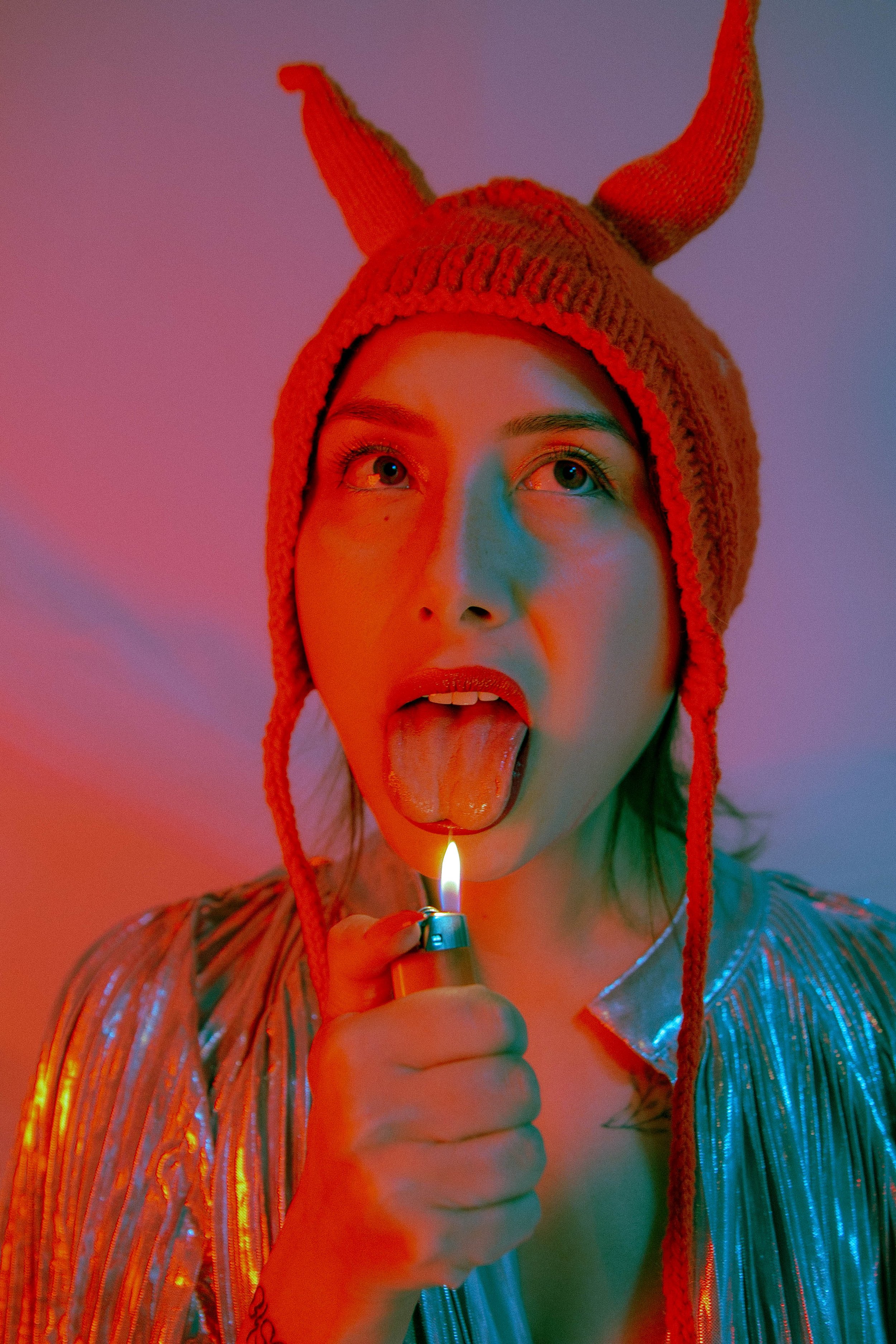 A woman with light skin wearing a red knitted hat with horns, holding a lighter to her tongue, with colorful lighting and a metallic striped top.