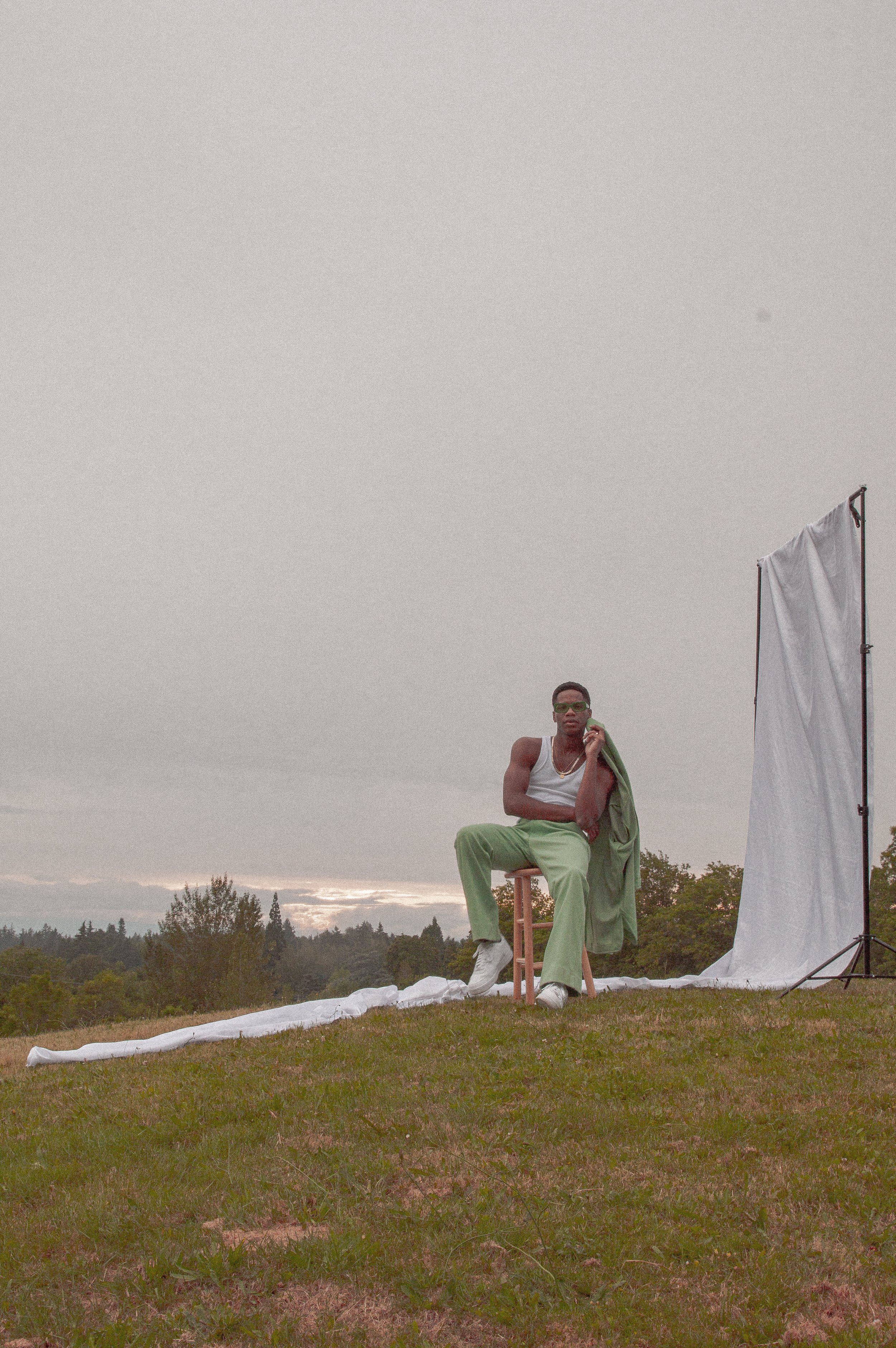 A man sitting on a stool outdoors, holding a phone to his ear with a cloth draped over his shoulder, against a cloudy sky and trees in the background. There is photography equipment and a white sheet or backdrop nearby.