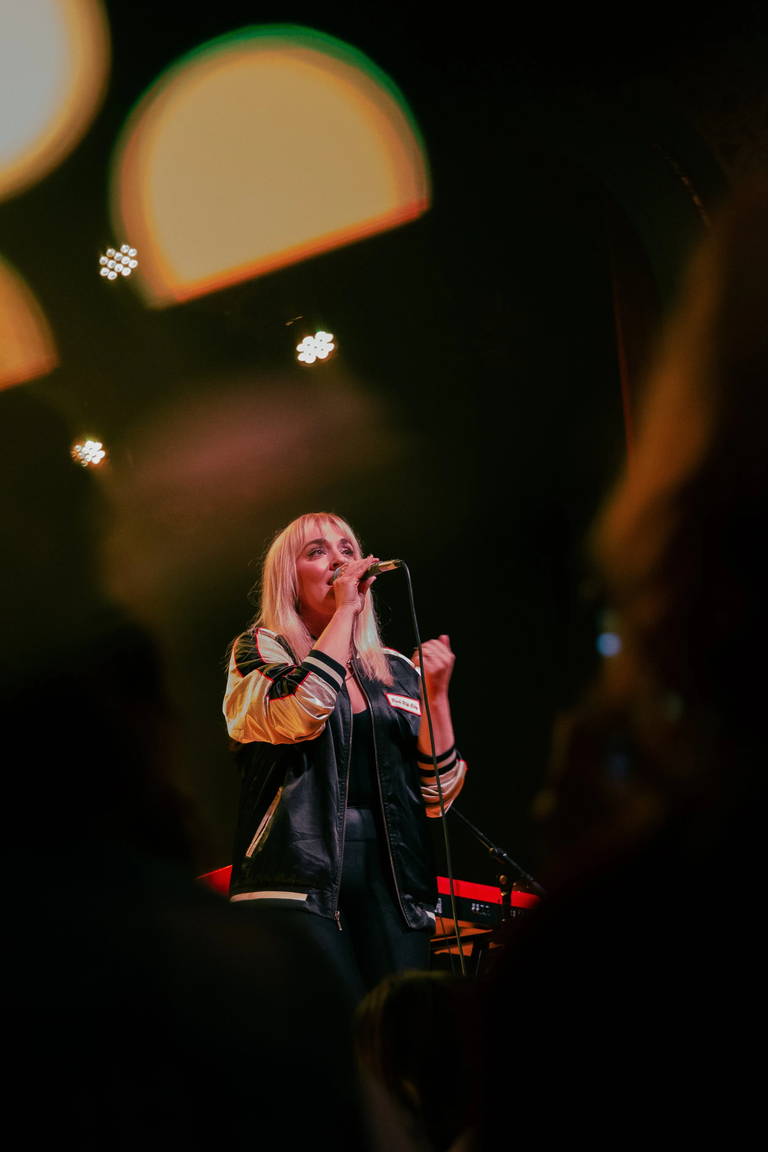 A female singer with blonde hair performing on stage, holding a microphone, with stage lights above her.