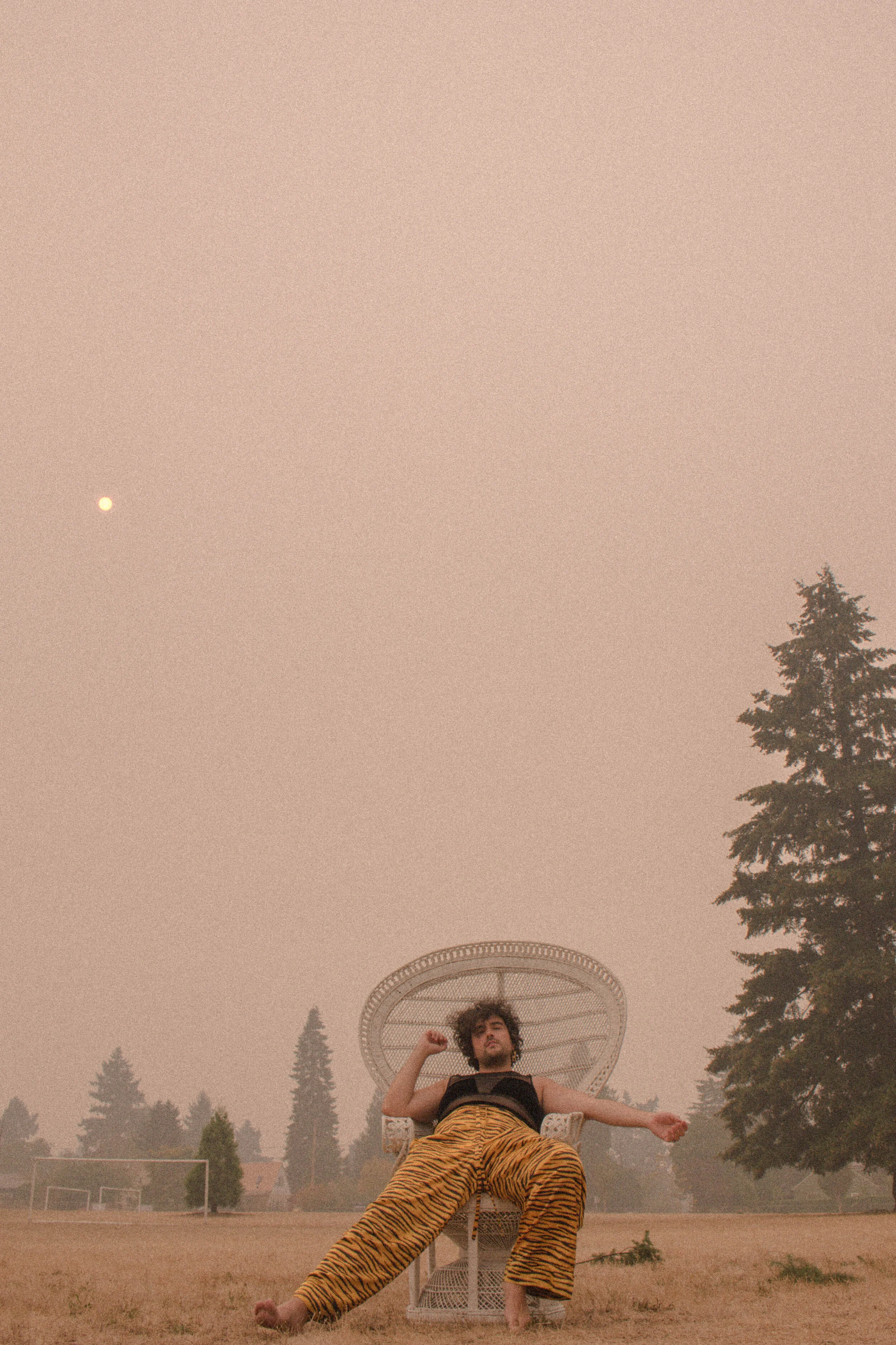 Person with curly hair wearing tiger print pants, sitting on a wicker chair outdoors in a park, raising fist, under a large circular structure, with trees, grassy field, sports field, and a partly visible moon in the sky.