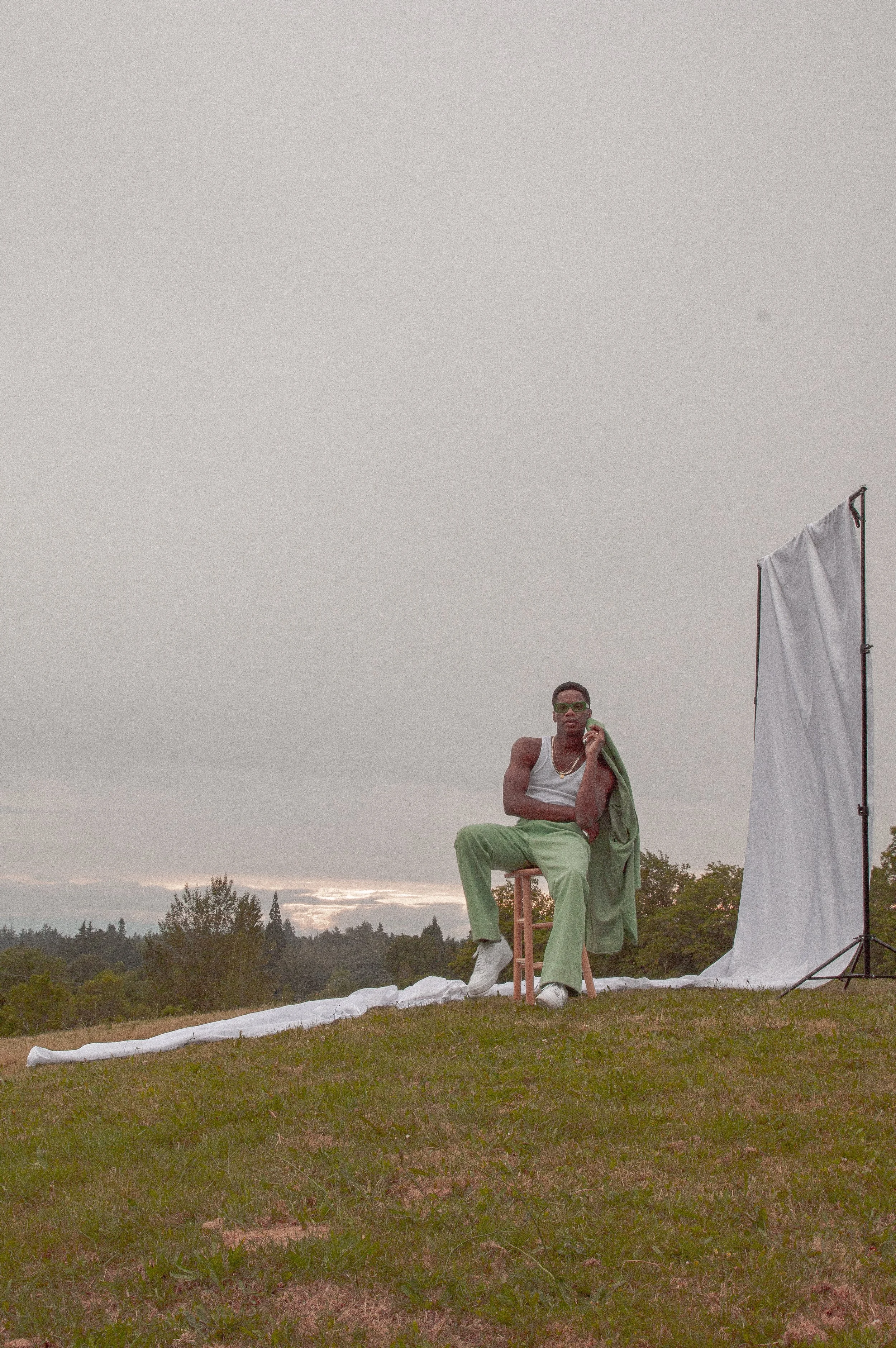A person wearing sunglasses and a white tank top sitting on a stool outdoors near a white backdrop and a flag, with trees and an overcast sky in the background.