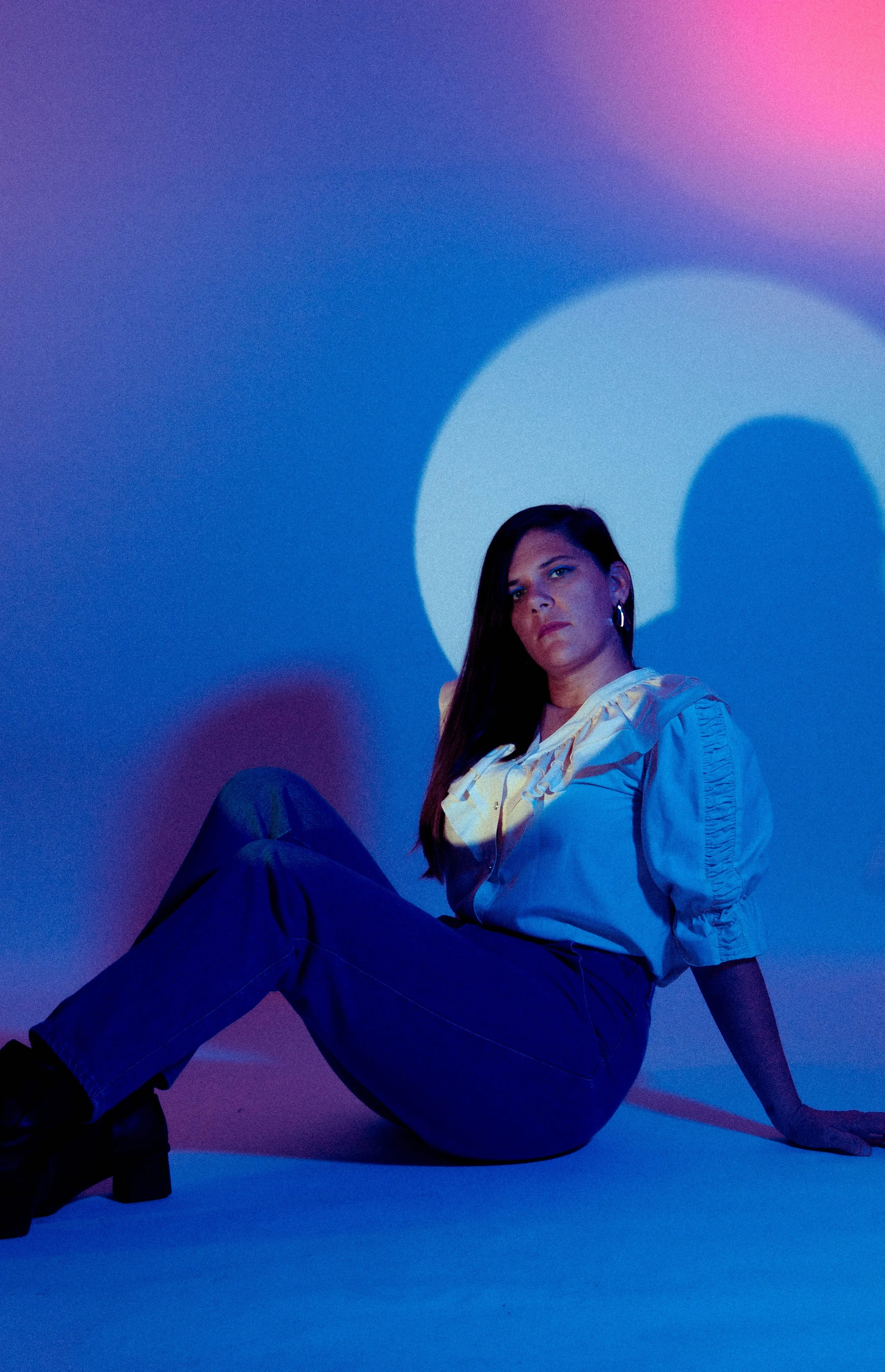 A woman sitting on the floor against a purple and pink gradient background, with large illuminated letter 'O' behind her. She is wearing a shiny, cream-colored blouse and dark pants, looking confidently at the camera.