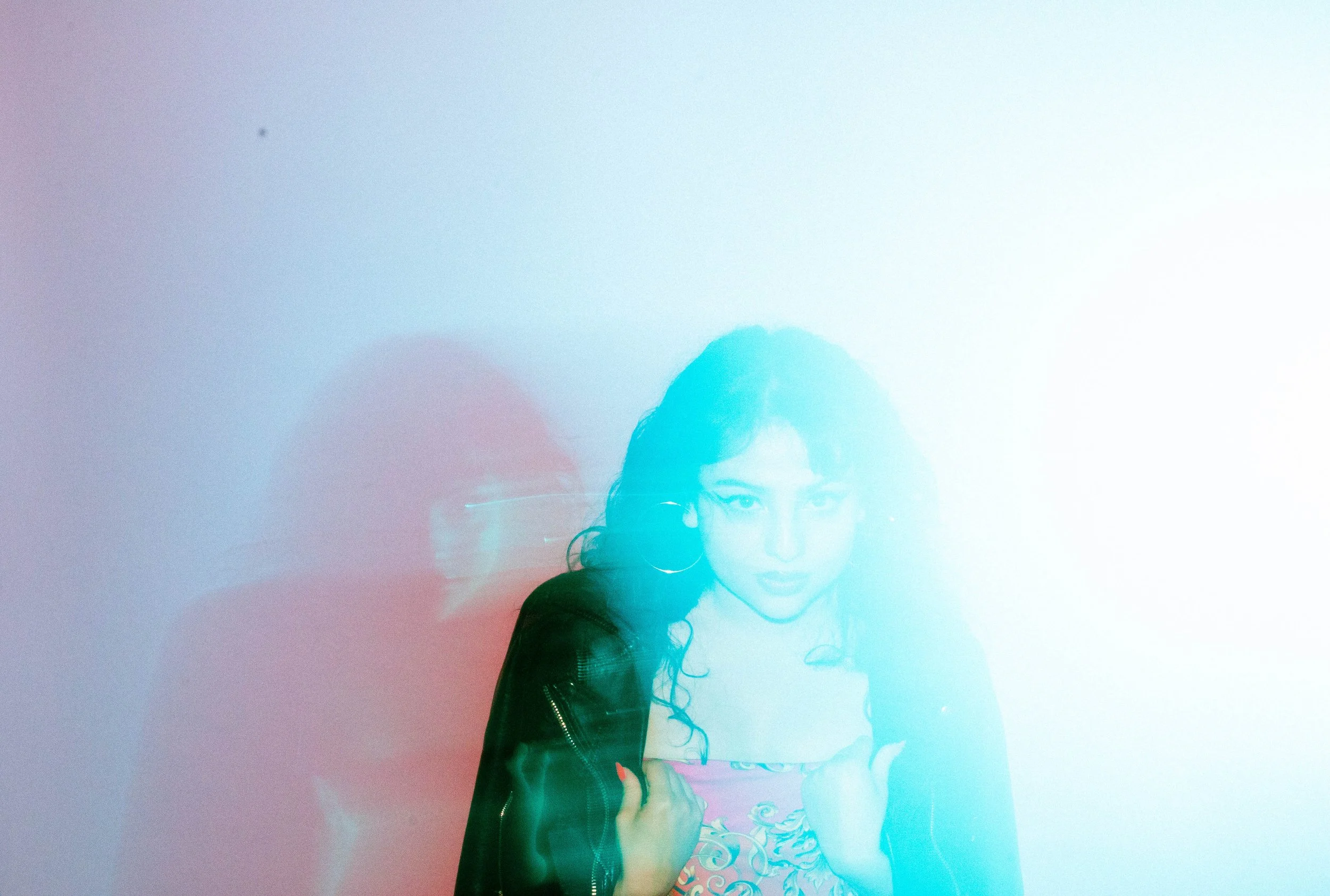 A woman with curly hair, wearing glasses and hoop earrings, holds a camera in front of a colorful, pastel gradient background.