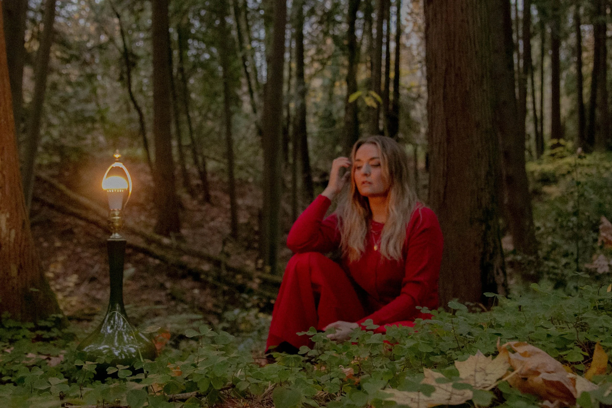 A woman with long blonde hair sitting in a forest at dusk, wearing a red jacket and red skirt, near a lit lantern on a small tripod.
