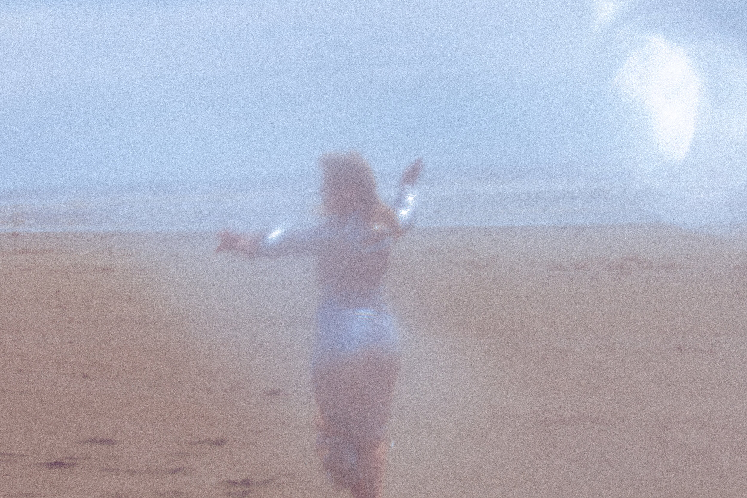 A person standing on a beach with a wide-open sky in the background, appearing to throw an object into the air, with a bright sun shining.