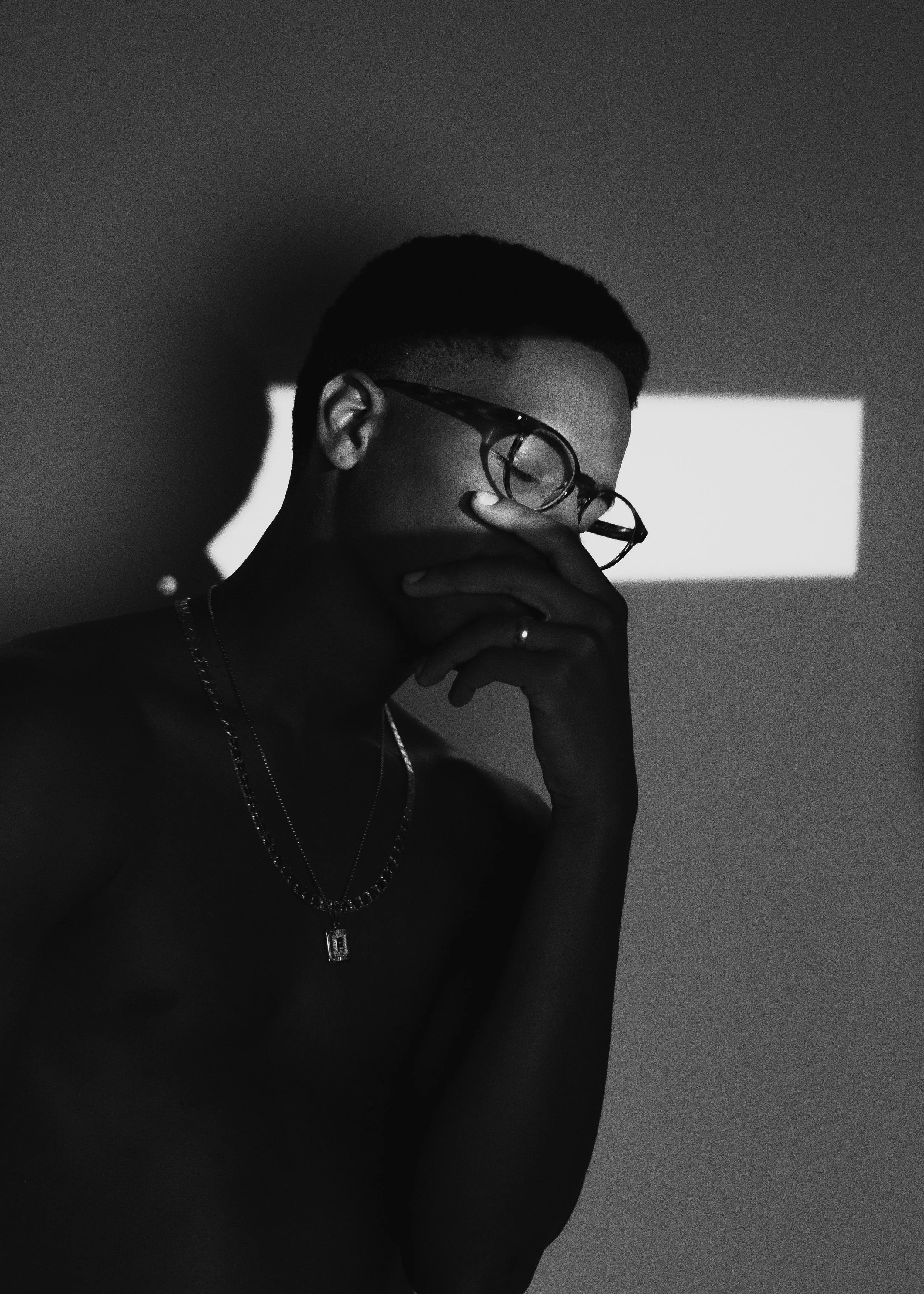 A black and white photo of a person with short hair wearing glasses, with their hand covering their mouth, in front of a background with a white rectangular light behind them.
