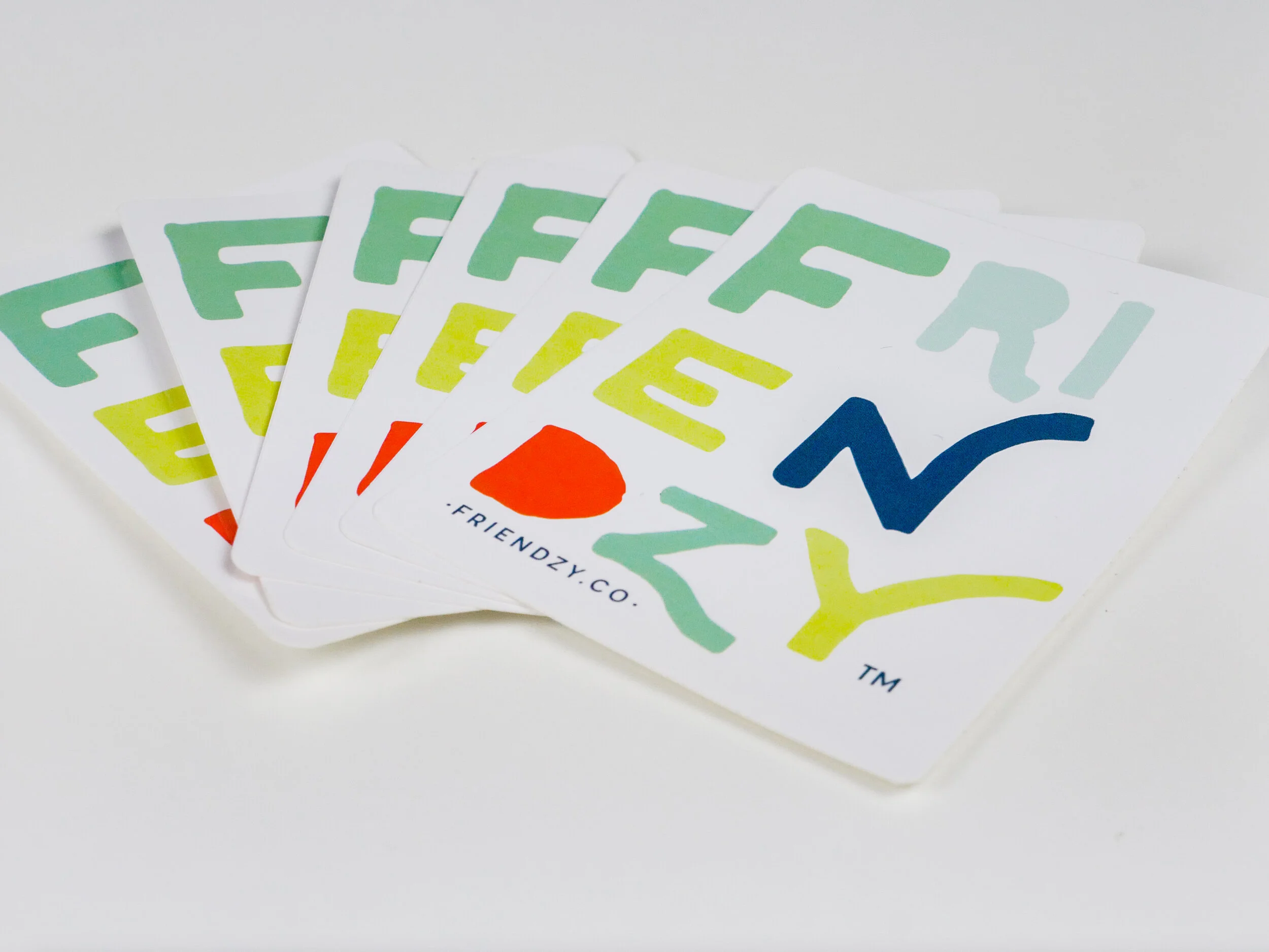 A set of colorful playing cards fanned out on a white surface, with cards featuring the F, I, R, E letters and the word 'Friendzy.co' at the bottom.