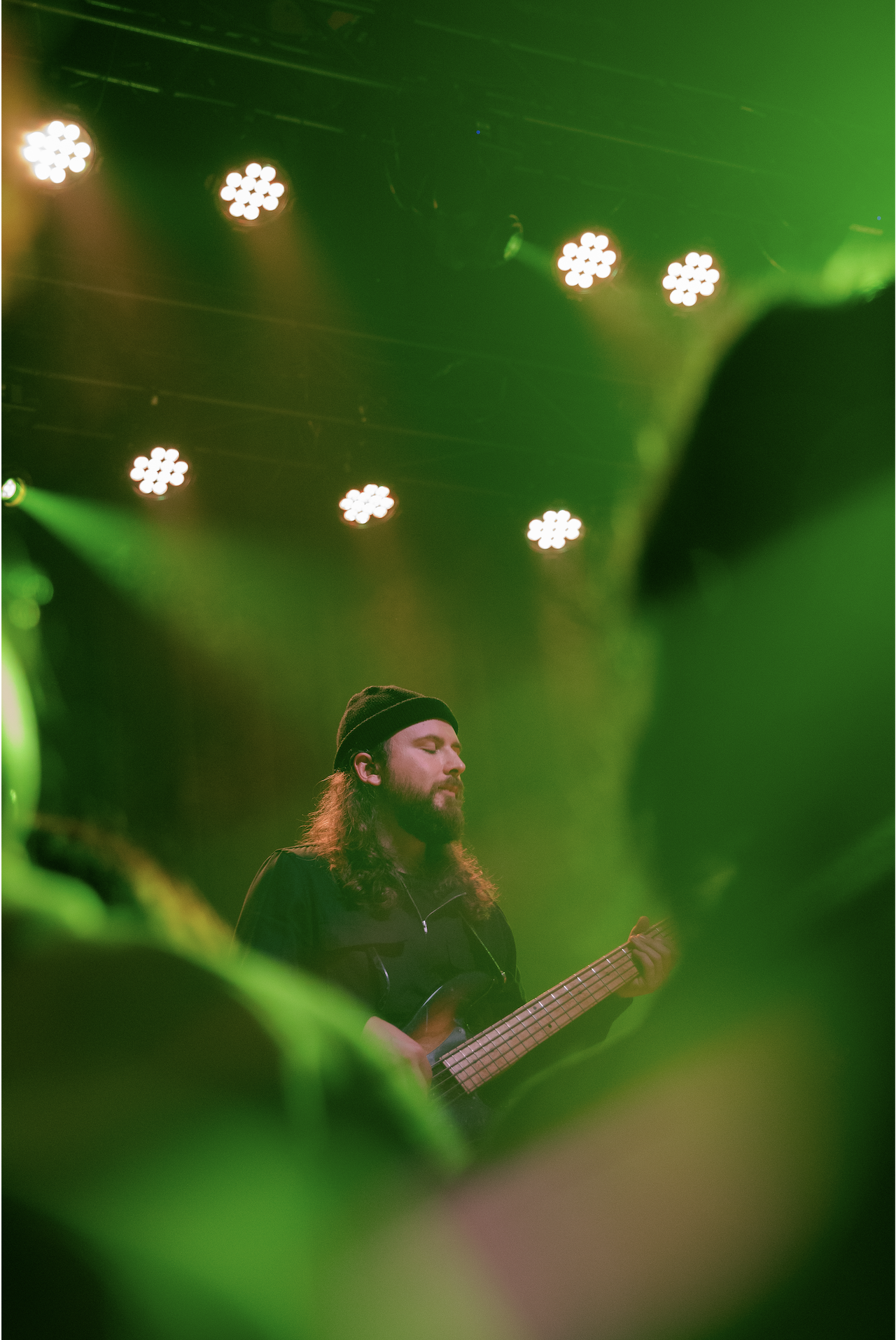 Musician with long hair and beard playing a guitar on stage, surrounded by green and yellow stage lighting, with spotlights overhead.