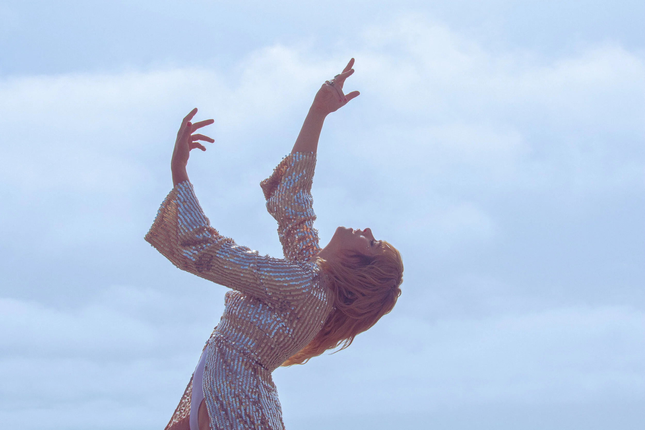 Person with long hair wearing a sequined jacket, raising their arms upward against a cloudy sky.