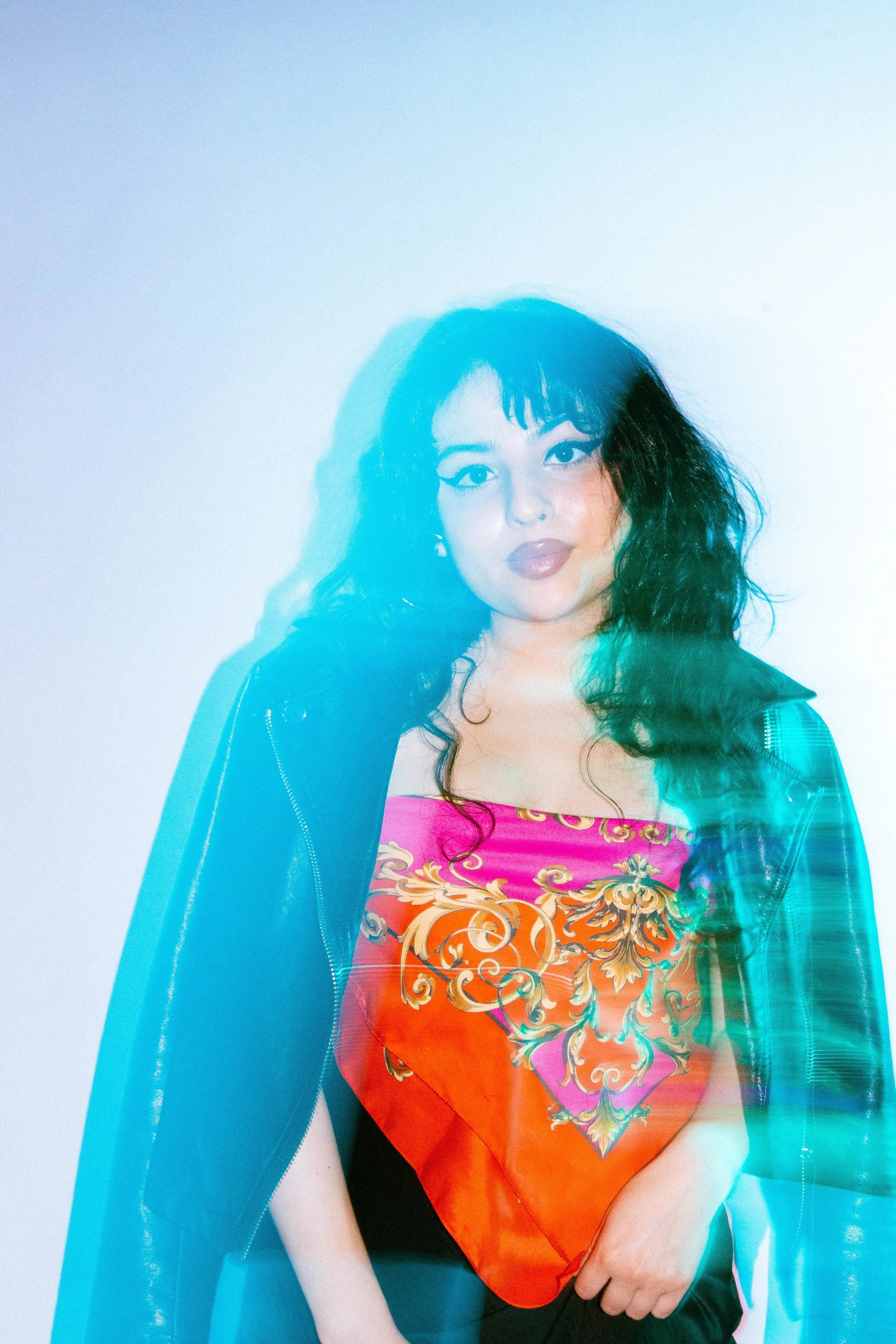 A woman with curly black hair and makeup wearing a black jacket over a colorful top with gold and floral patterns, against a light blue background with a layered effect.