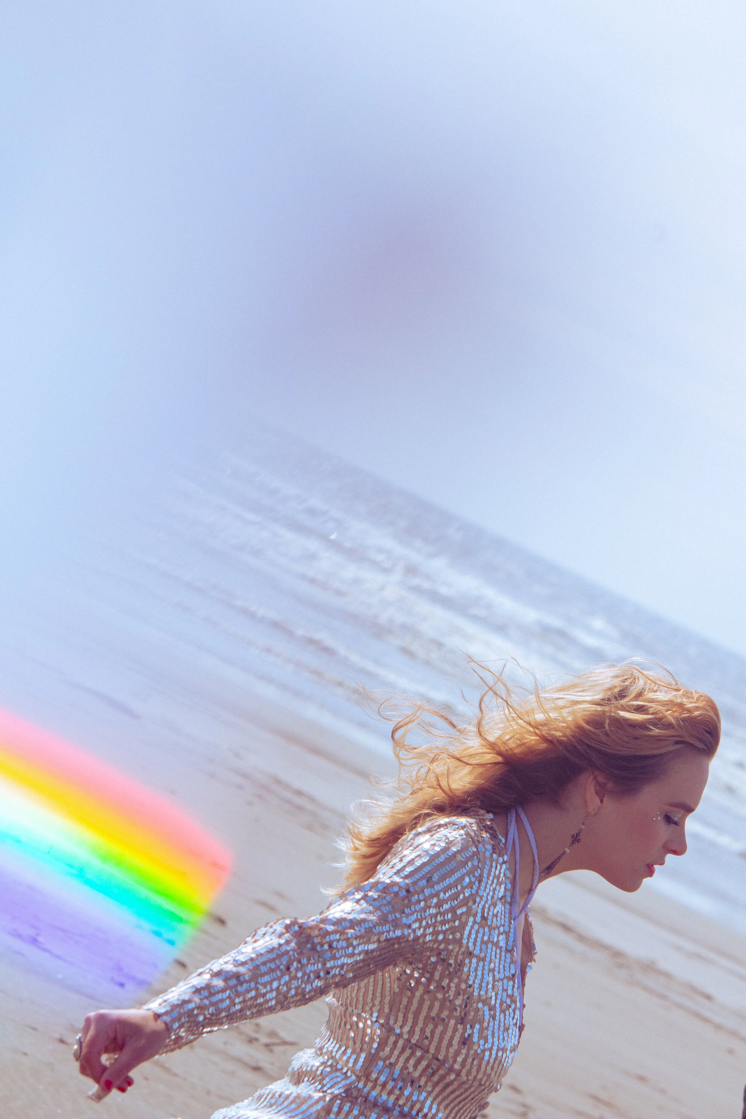 A woman with curly blonde hair running along the beach with the ocean and sky in the background. She is wearing a shimmery, striped dress and appears joyful.
