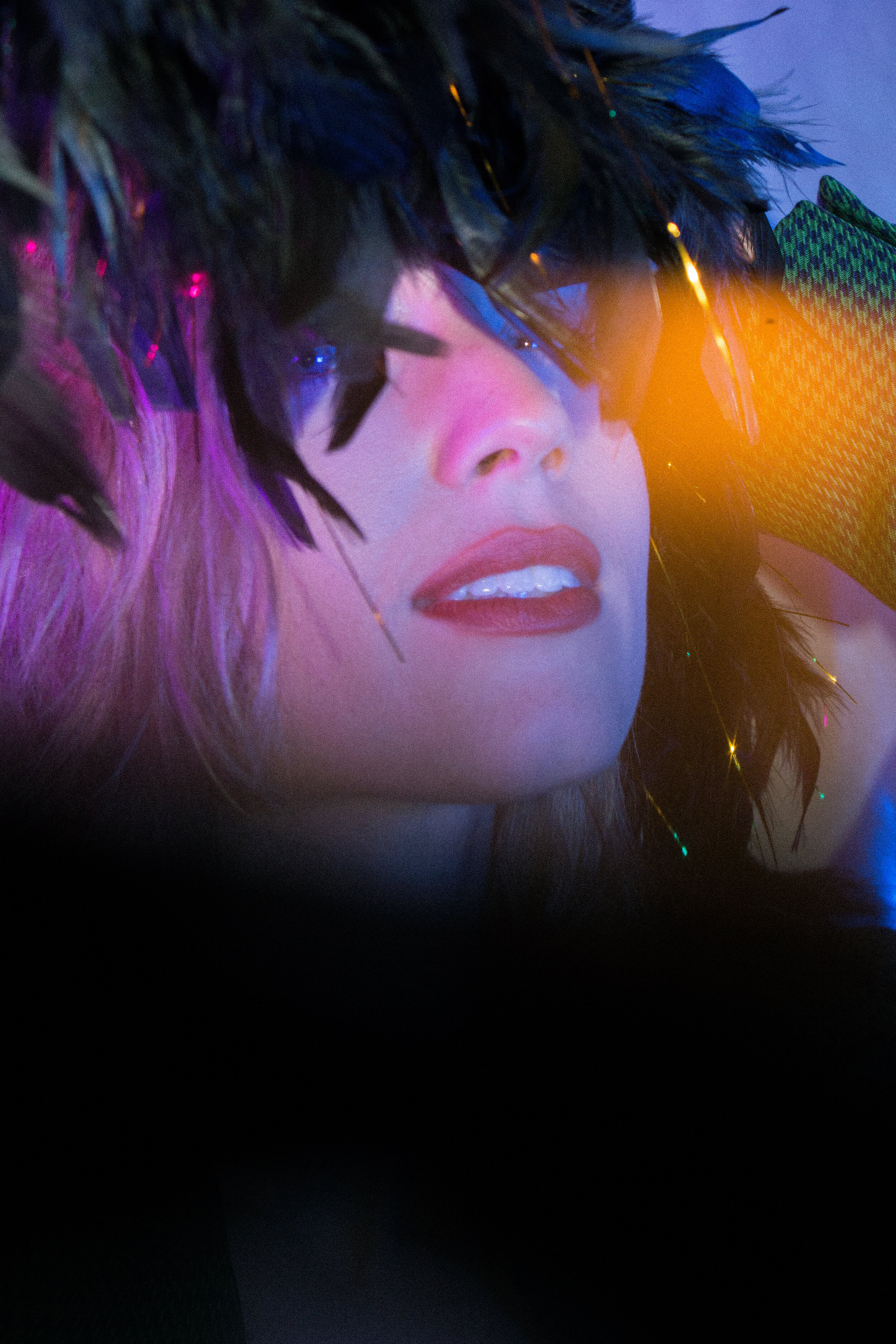 Close-up of a person's face with makeup, pink lipstick, and dark eye makeup, partially covered by colorful feathers with some of the feathers reflecting colored lights in a dark setting.