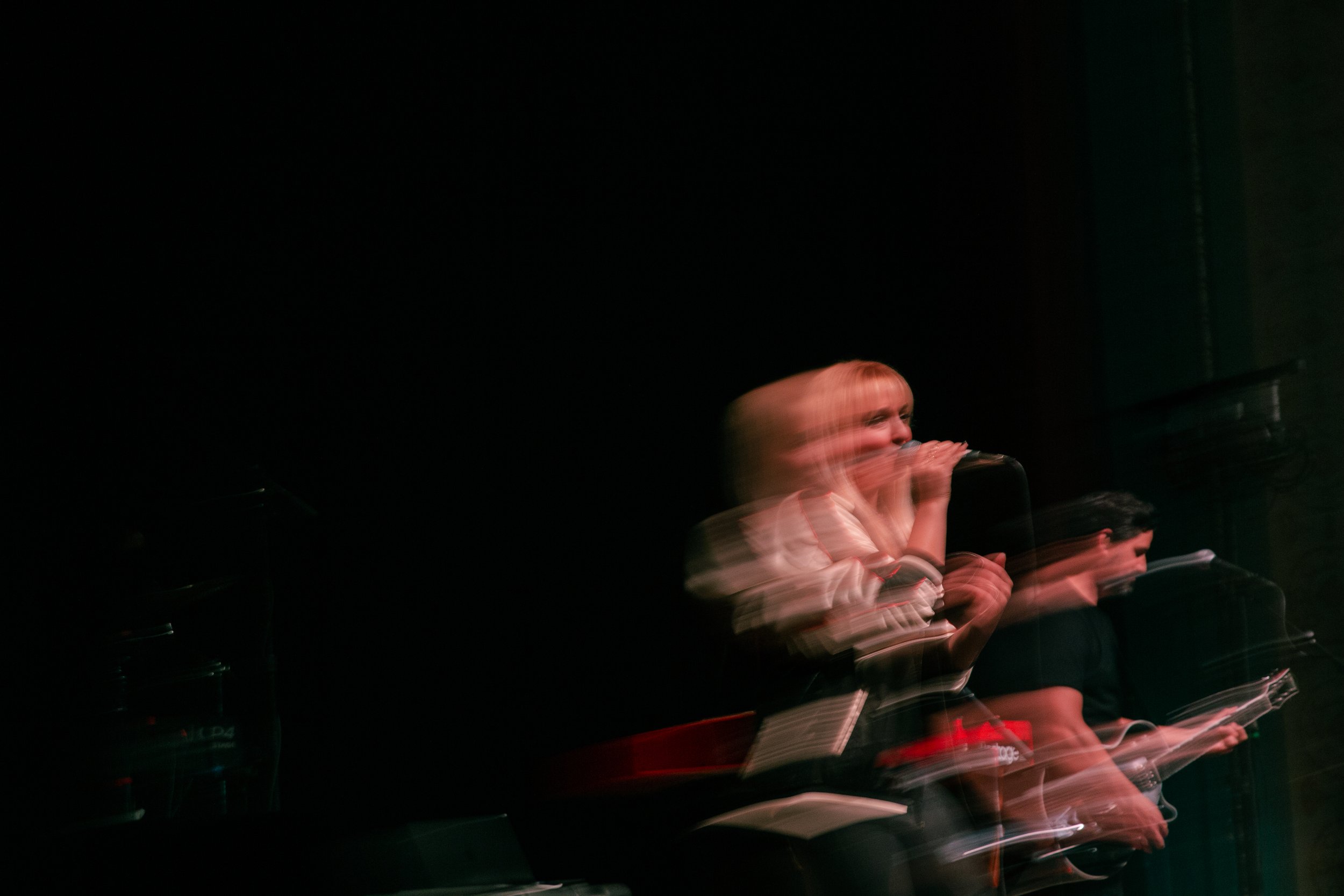 A person with blonde hair singing or speaking into a microphone, with motion blur indicating movement, in a dark environment.