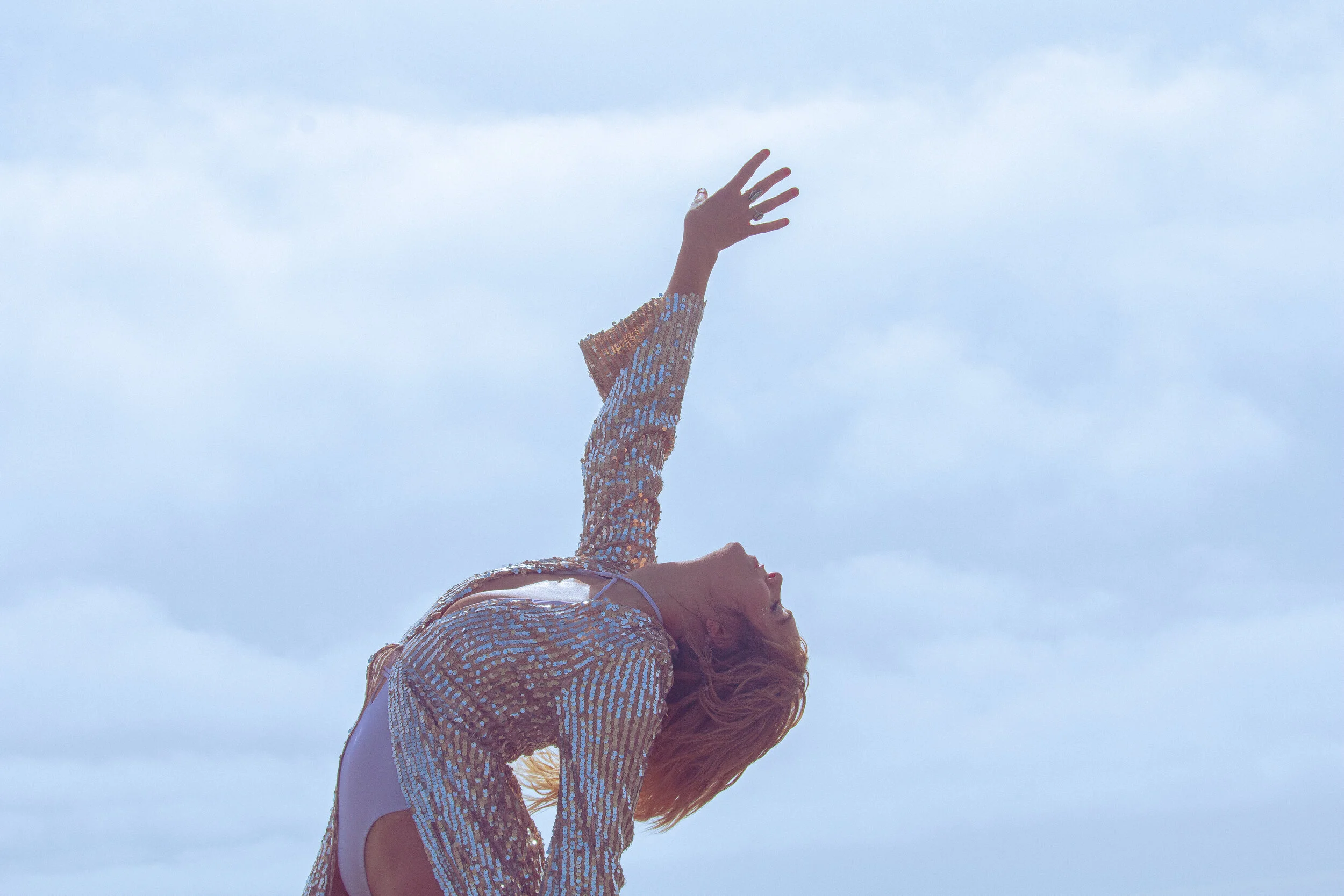 A woman with long red hair is lying on her back outdoors, wearing a shiny, sequined top, reaching her right arm upward toward the cloudy sky.