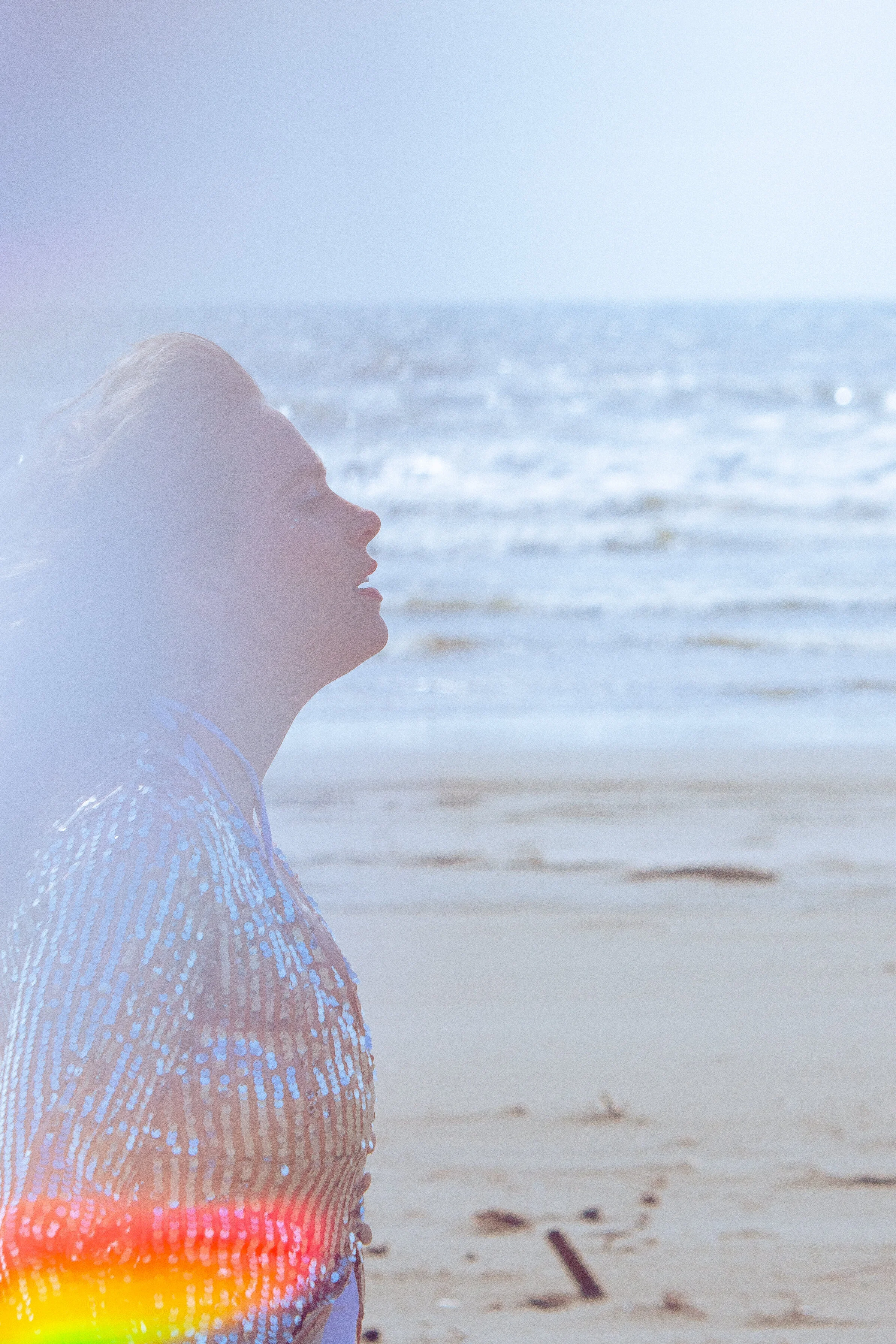 A woman with brown hair at the beach, facing right, with waves in the background and a rainbow-colored light effect.
