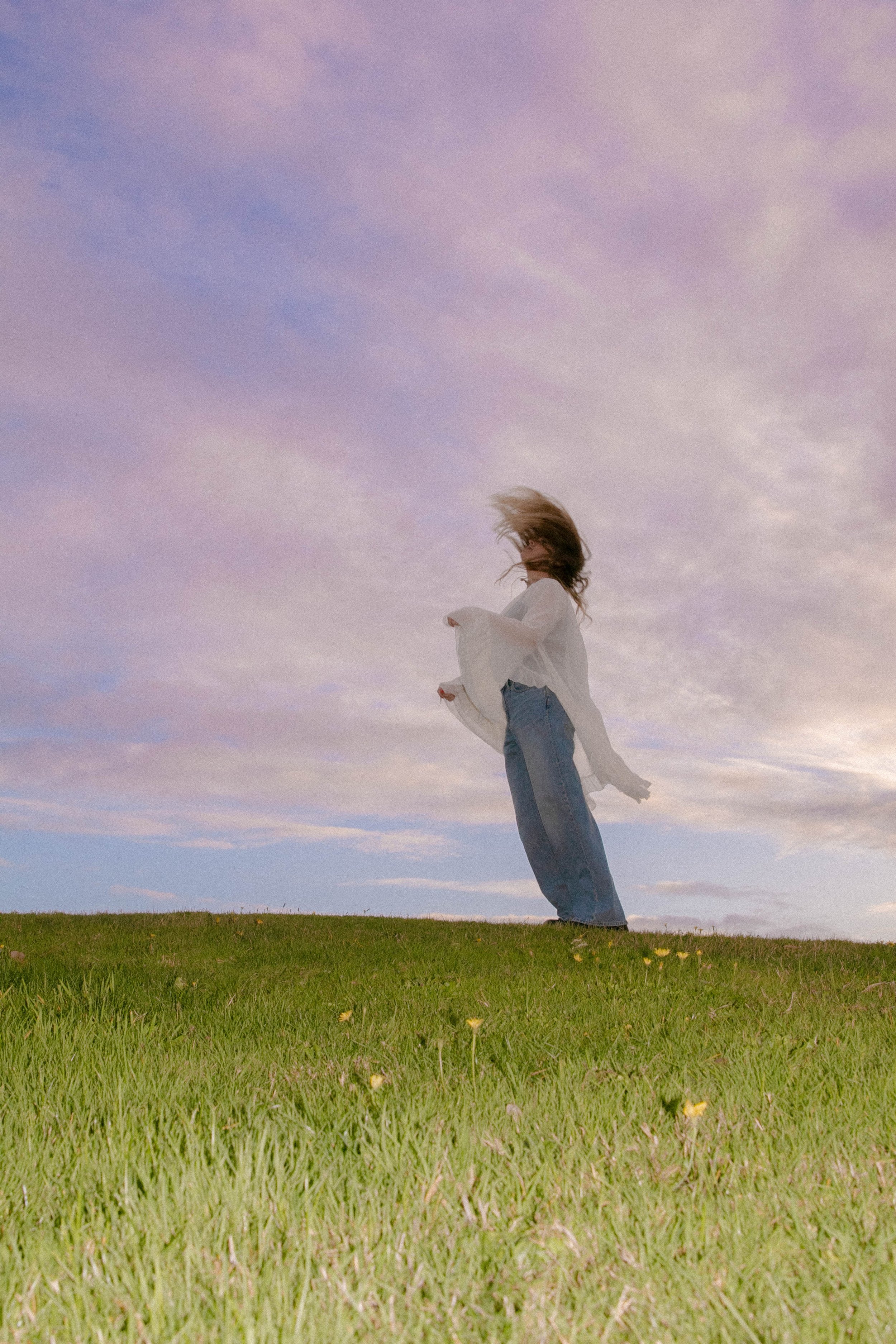 A person with long hair blowing in the wind, standing on a grassy field with a cloudy sky in the background.