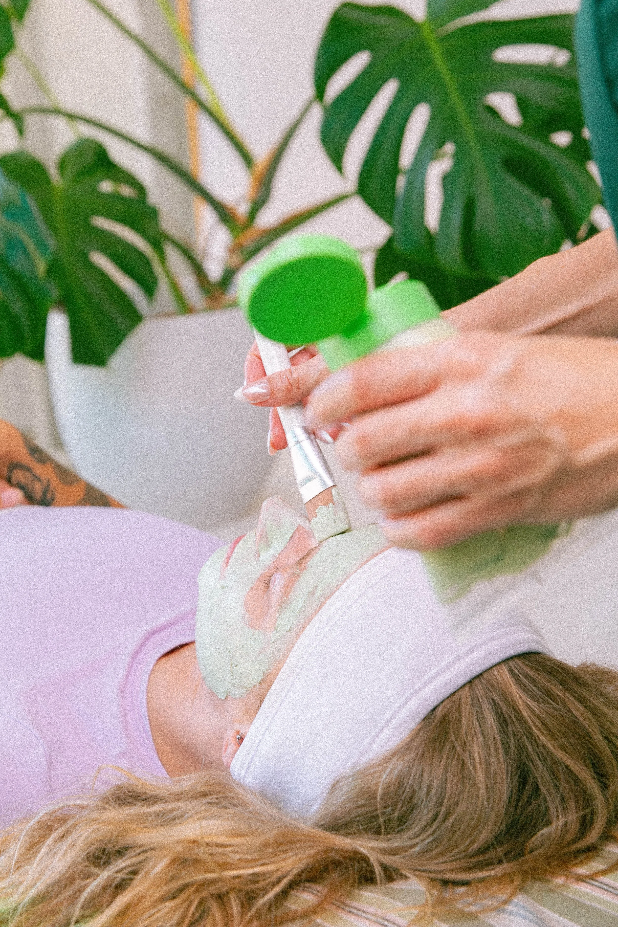 A woman lying on her back with a facial mask applied to her face, receiving a skincare treatment with a green silicone brush at a spa or skincare clinic, with green plant leaves in the background.