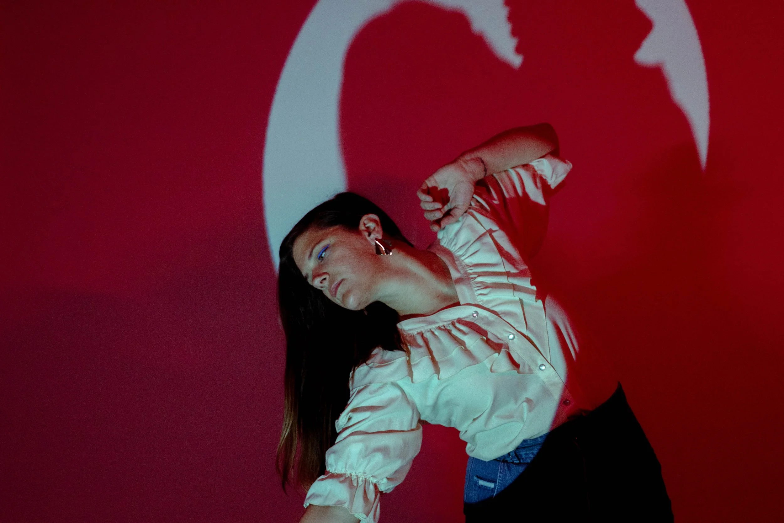 A woman with long dark hair, wearing a cream-colored shirt with ruffled sleeves, is resting her head on her arm against a red background. Shadows of large, stylized red hearts are projected on the wall behind her.