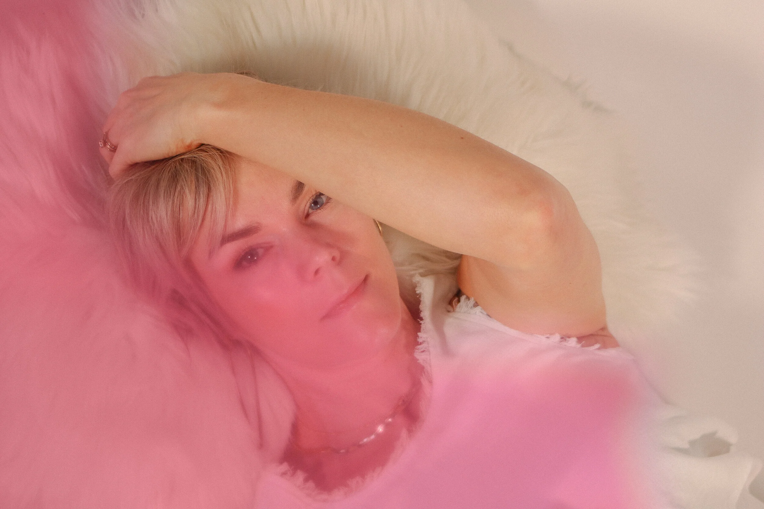 A woman with blonde hair lying on a pink fluffy blanket, resting her head on her arm, looking at the camera.