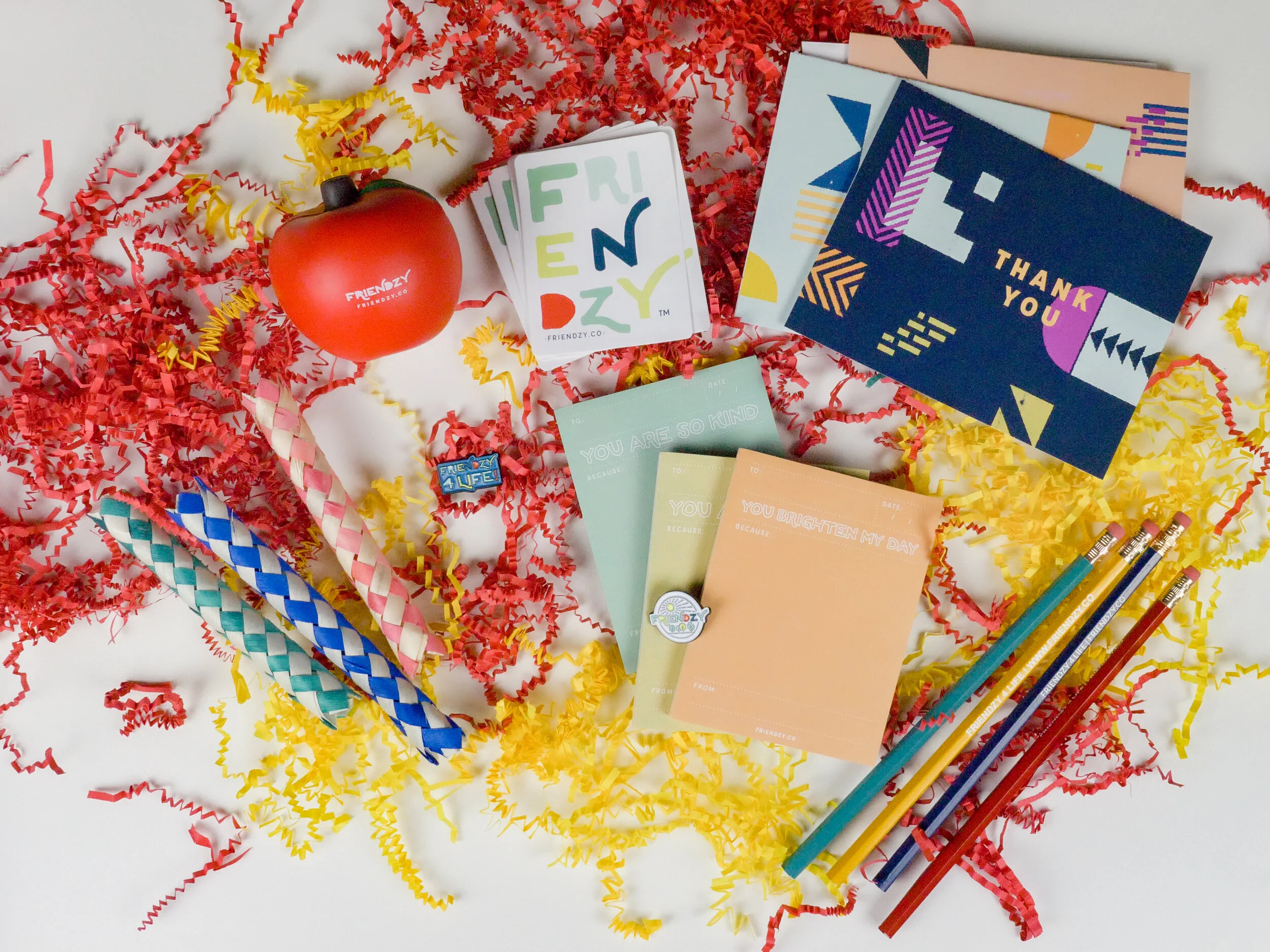 Colorful celebration items on a white surface, including a red balloon with 'Friendly' written on it, a small stack of greeting cards with bright, written messages, striped paper straws, and yellow and red shredded paper confetti.