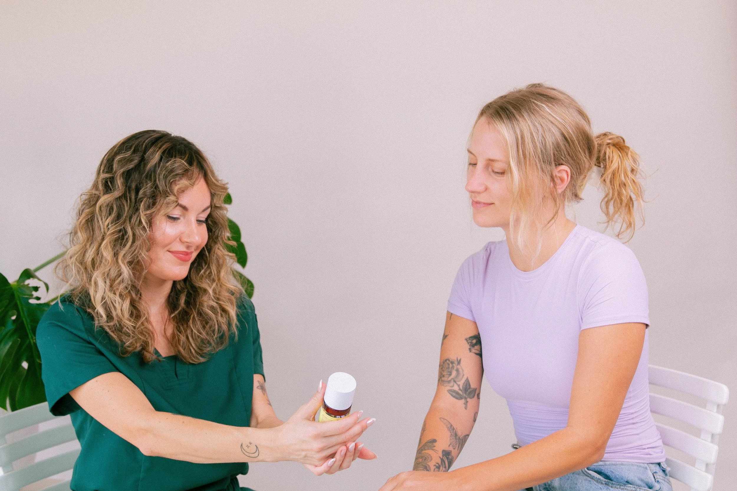 A woman in medical scrubs holding and showing a bottle to a woman with tattoos on her arms, sitting indoors with a white wall background.