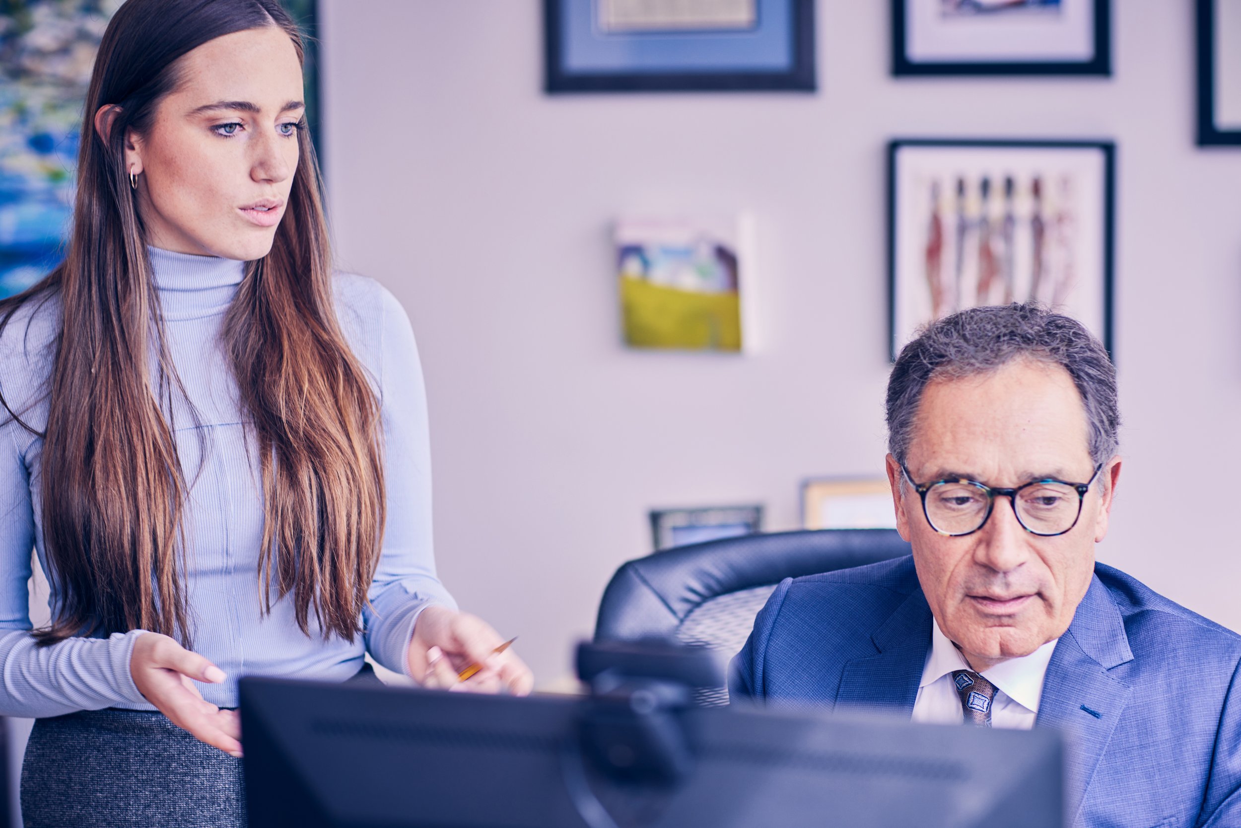 A young woman with long brown hair, wearing a light blue blouse, is standing next to an older man with glasses and gray hair, who is seated at a desk working on a computer. They are in an office with framed pictures on the wall.