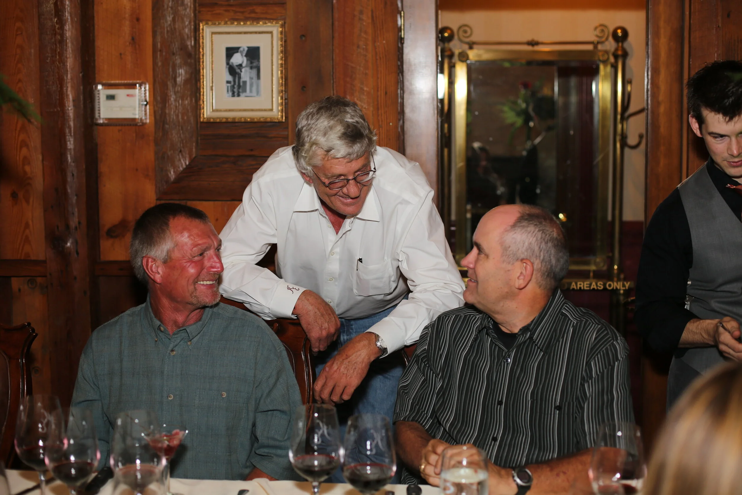 Three men interacting at a dining table with wine glasses, in a wooden-paneled room with framed pictures on the wall.