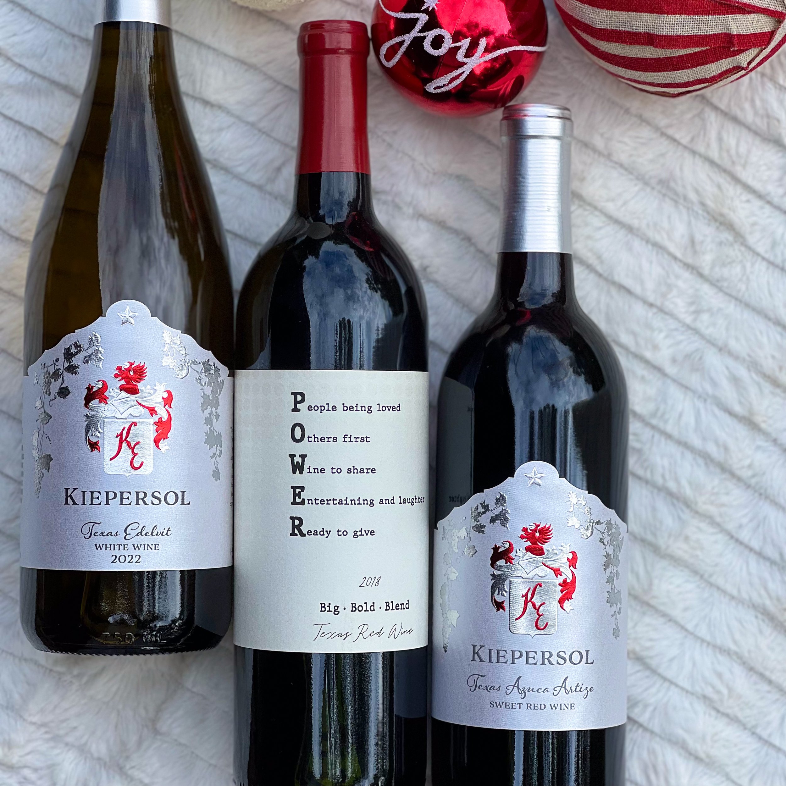 Three bottles of Kiepersol wine on a white textured surface. The bottles include a 2022 Texas Edelzct White Wine, a 2018 Big Bold Blend Texas Red Wine labeled POWER, and a Texas Sweet Red Wine. Red and white decorative ornaments are partially visible