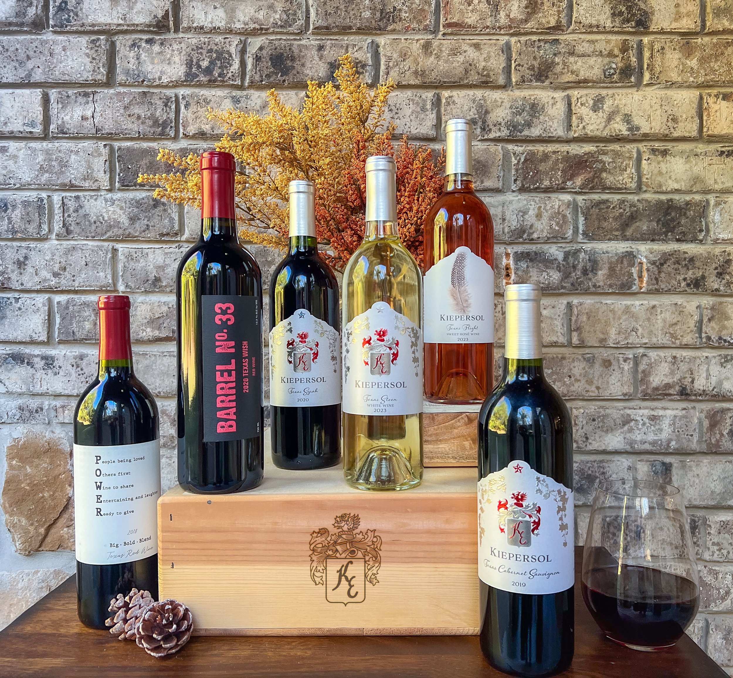 Assorted wine bottles displayed on a wooden crate, with a brick wall backdrop and decorative dried plants.