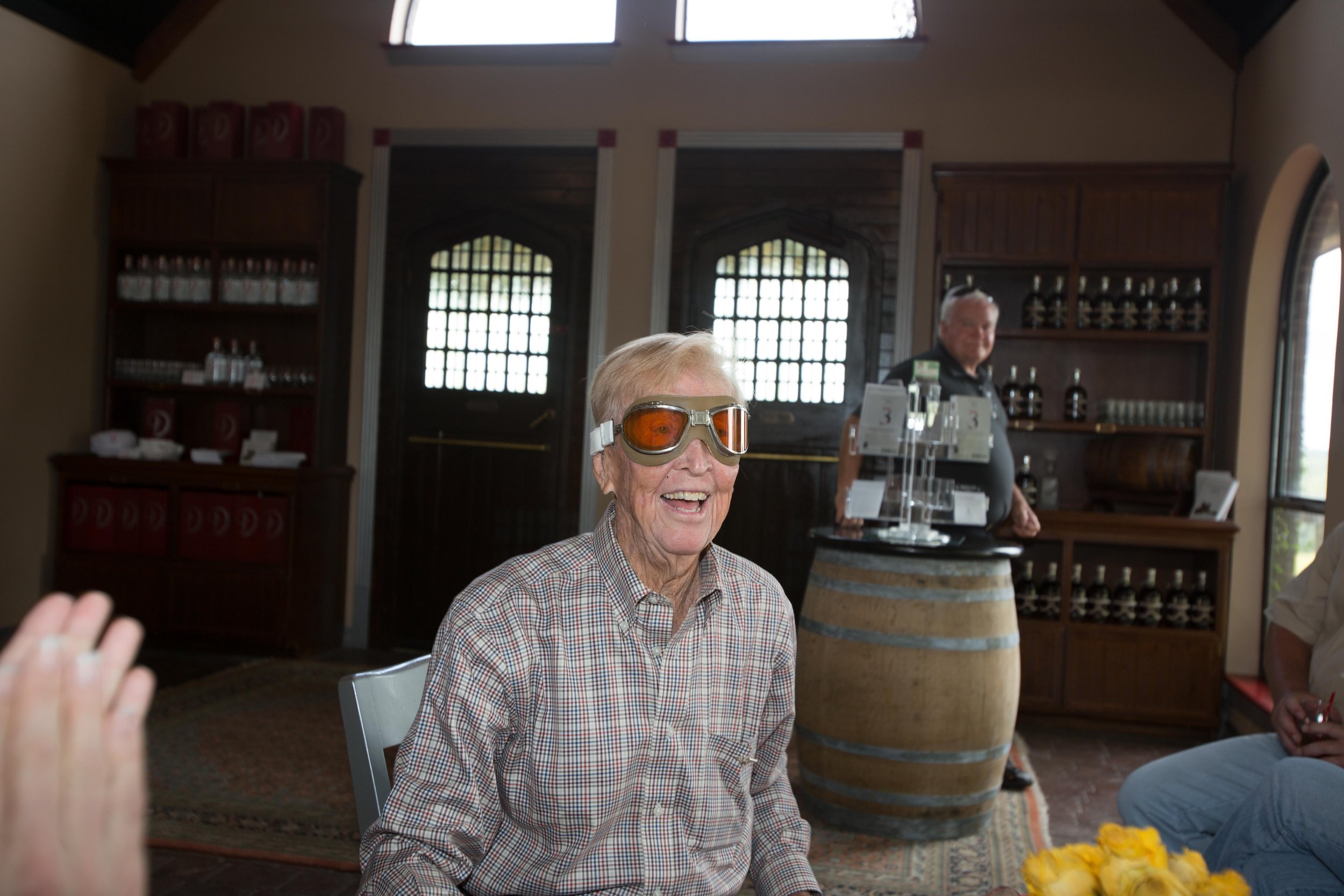 An elderly man wearing ski goggles, smiling indoors. Behind him is a room with shelves of bottles and a man standing by a barrel.