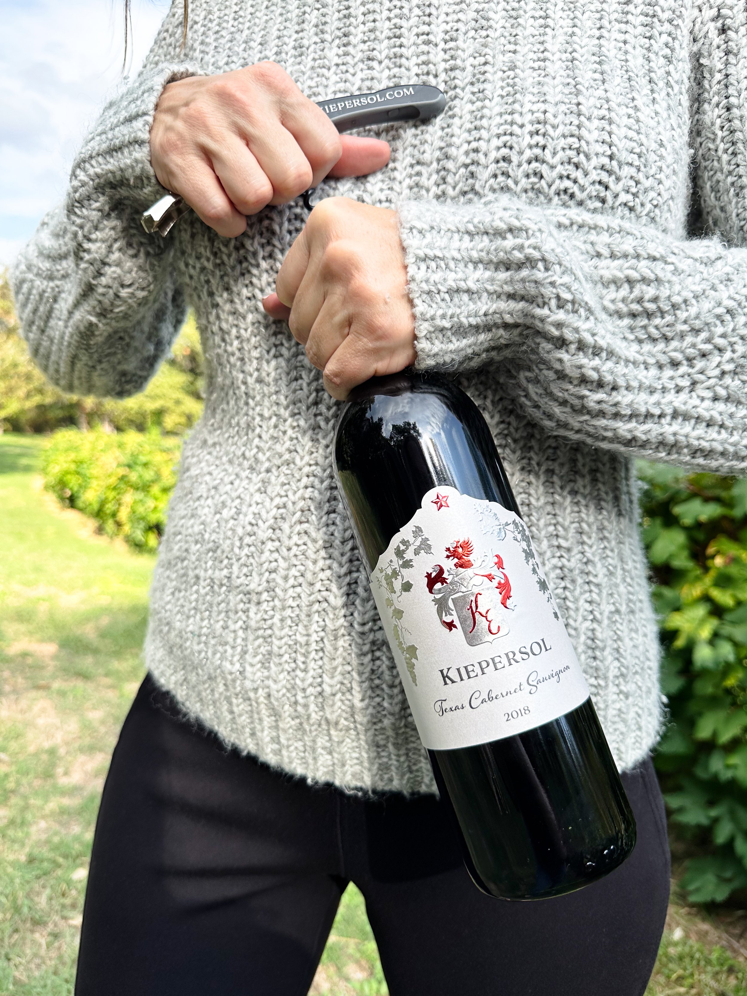 Person holding a bottle of Kiepersol Texas Cabernet Sauvignon 2018 with a corkscrew outdoors.