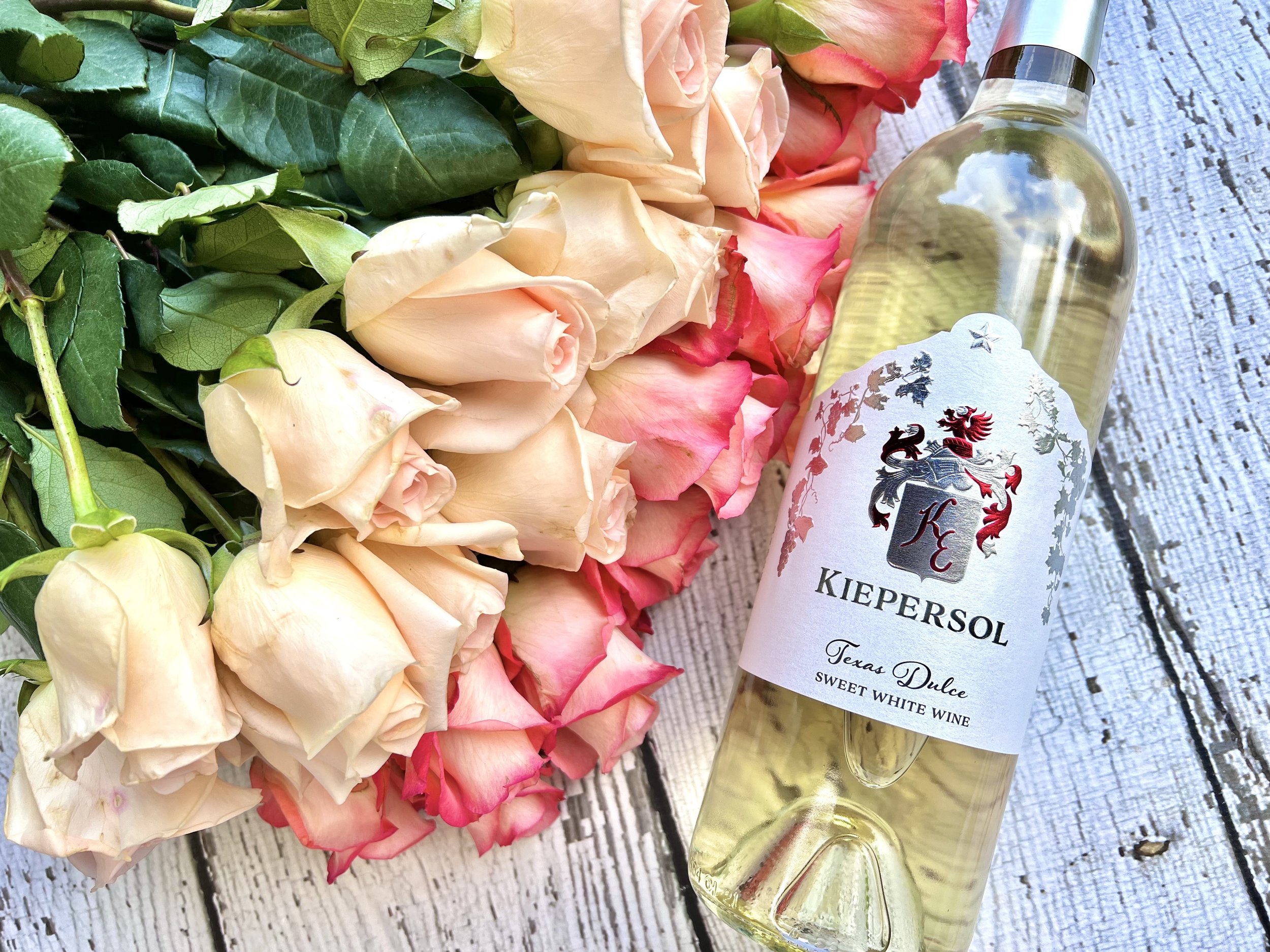 A bottle of Kiepersol sweet white wine labeled 'Texas Dulce' next to a bouquet of pink roses on a wooden surface.