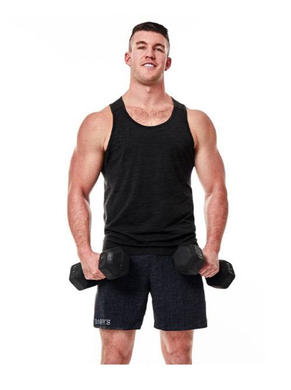 A man standing and holding two dumbbells, wearing a black tank top and dark shorts, smiling at the camera.