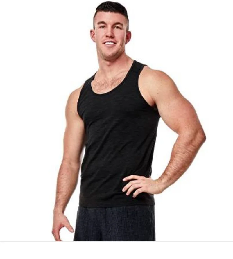 A young man with short dark hair, wearing a black tank top and dark shorts, smiling and posing with one hand on his hip.