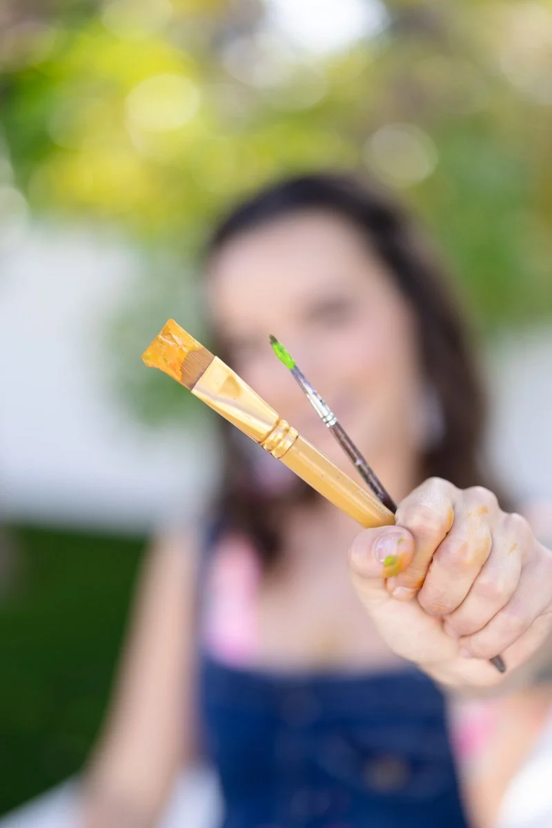 A woman holding two paintbrushes towards the camera, one with orange paint and the other with green paint, with a blurred background and smiling face.