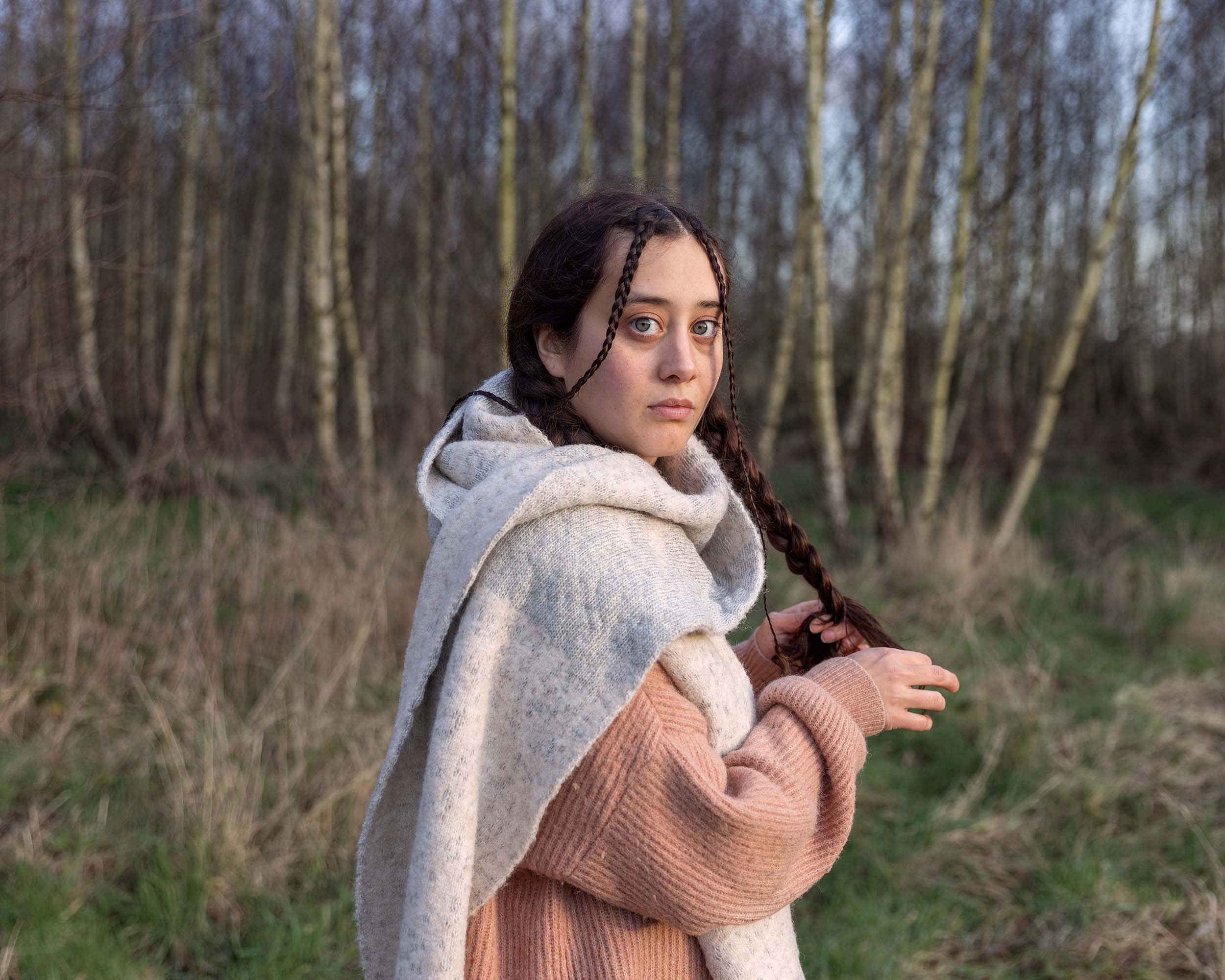Portrait of a girl with braided hair in a forest wearing comfortable winter clothes. She's wearing a fluffy pastel pink jumper and a bulky knit scarf. Image by Bieke Depoorter