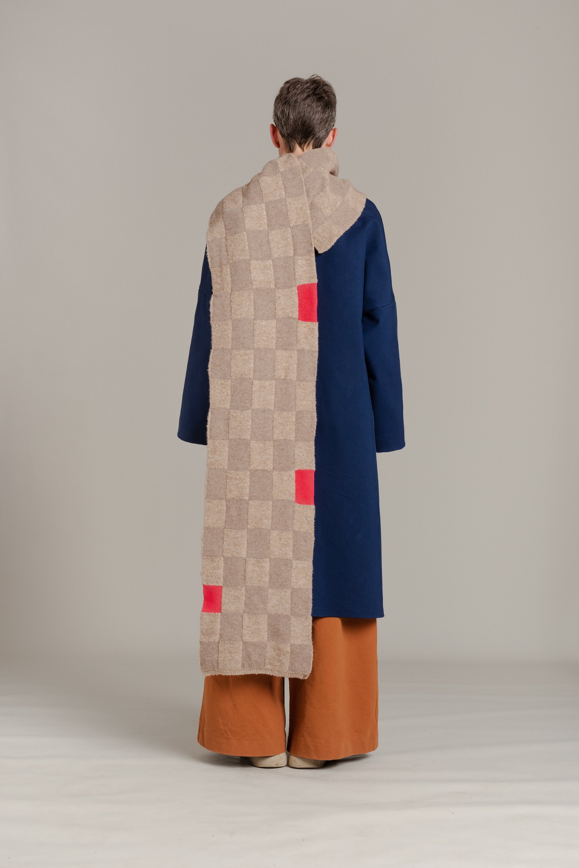 Wide shot of a woman with a knitted scarf draped over her back. She's wearing a dark blue winter coat and wide leg trousers. The scarf has a checkered pattern in beige with coral pink details.