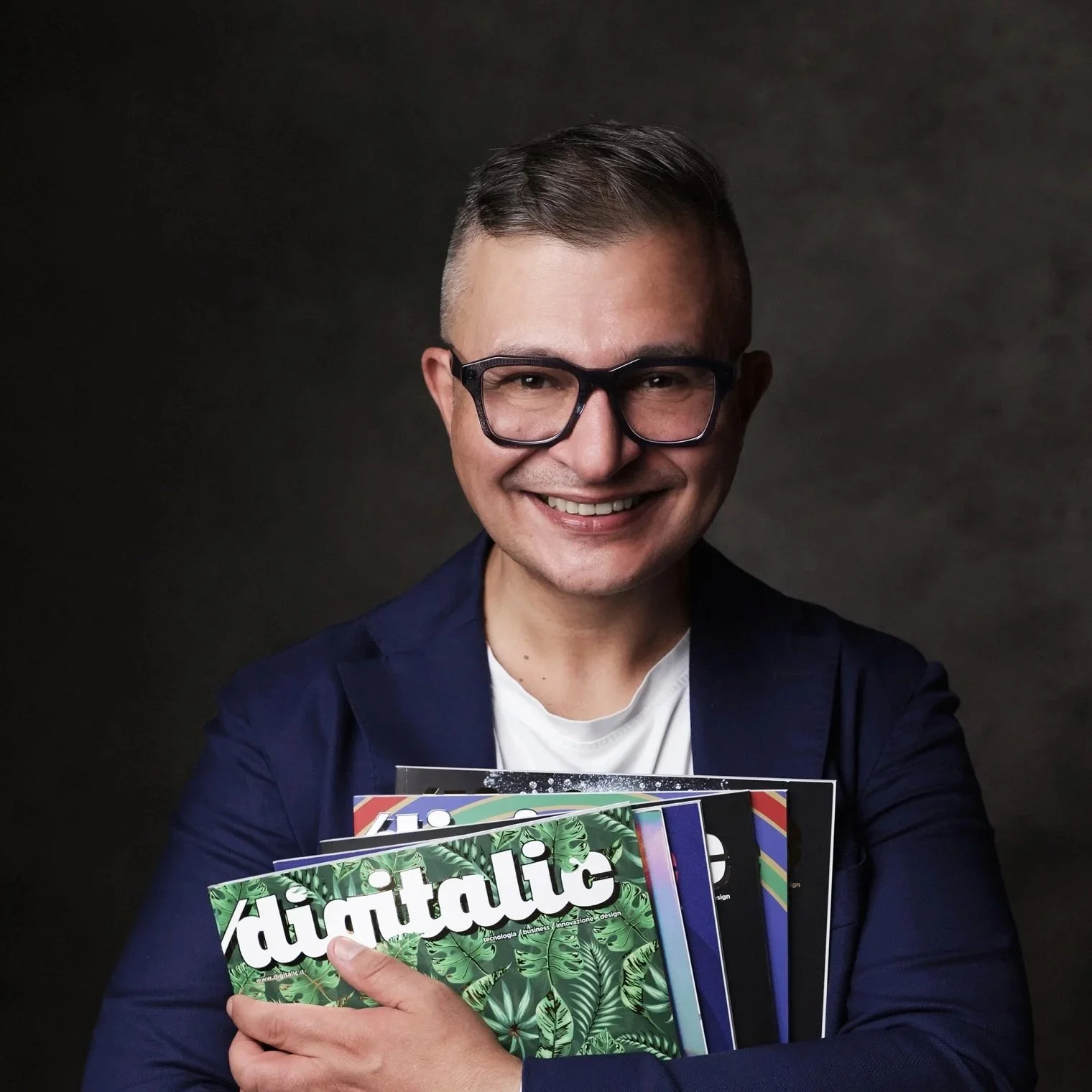Uomo sorridente con capelli scuri, barba leggera, indossa giacca blu e maglietta celeste, sfondo con piante verdi e finestra.