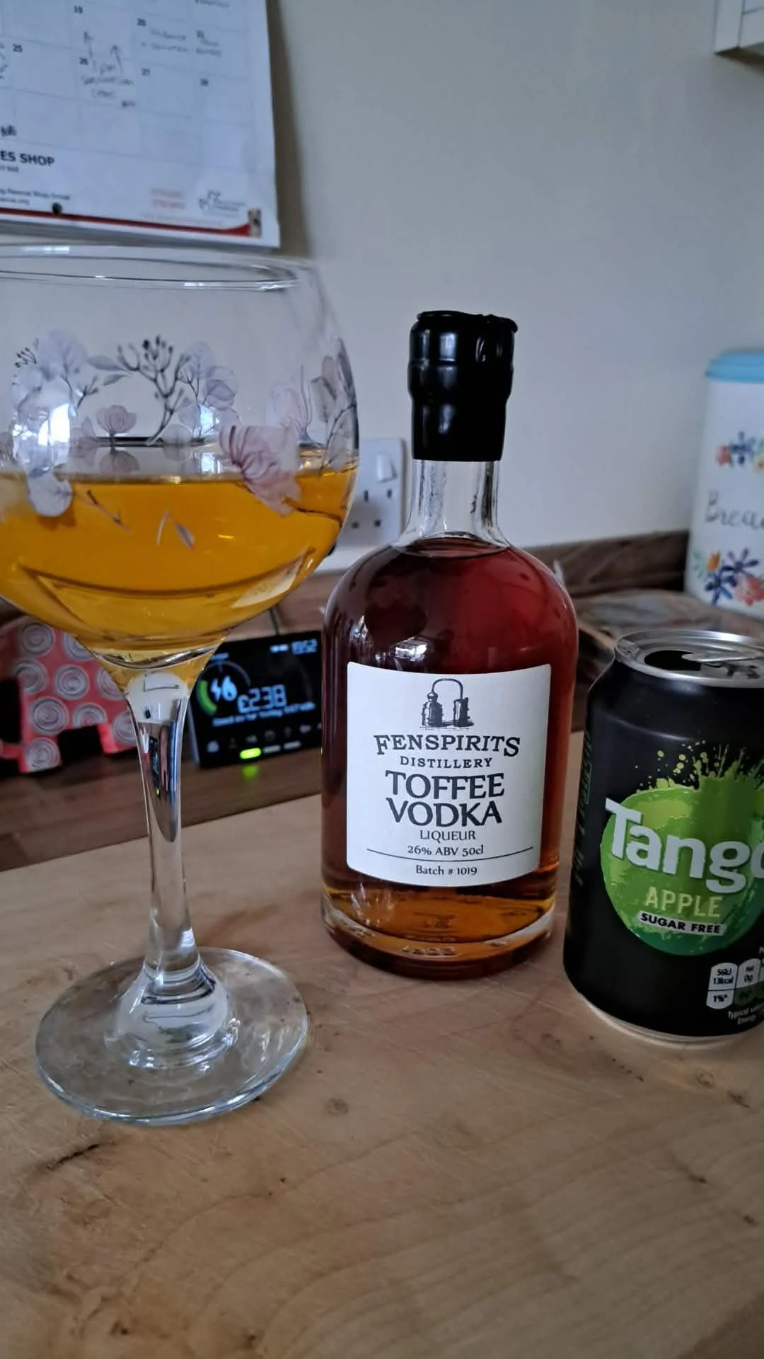 A glass with a floral design filled with a yellow-orange beverage, a bottle of Fenspirits Toffee Vodka, a can of Tangy Apple sugar-free drink, in a kitchen setting with a calendar and electronic device visible in the background.