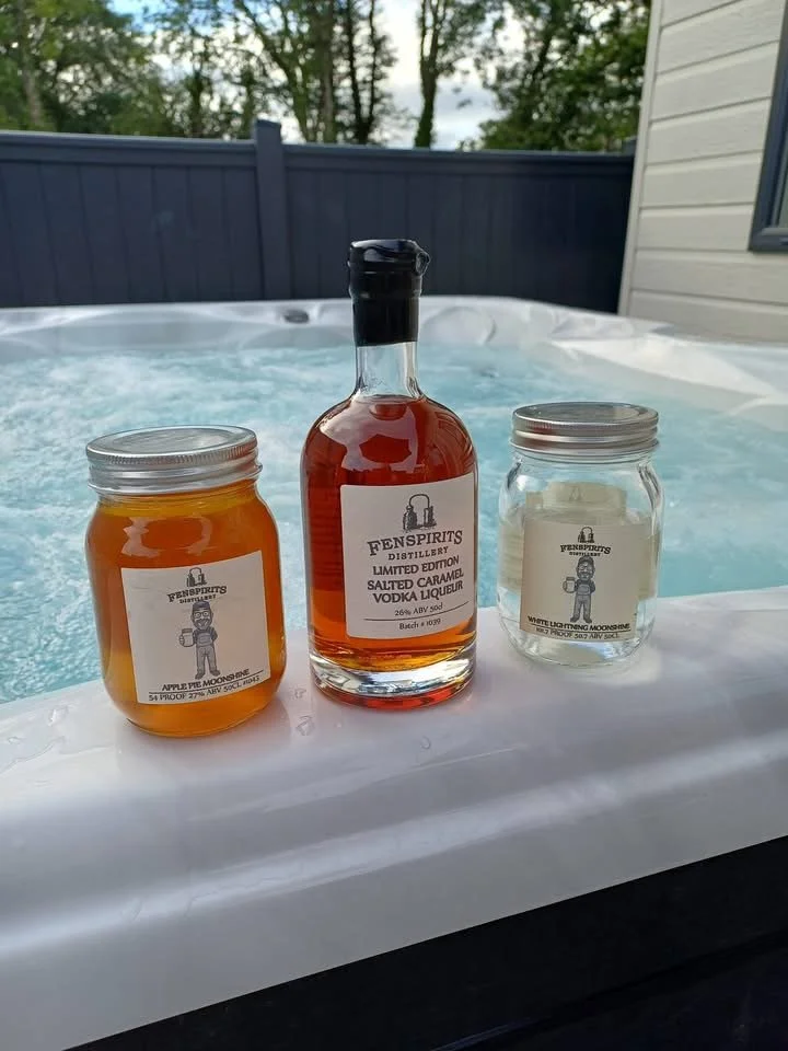 Three jars and a bottle on the edge of a hot tub. The jar on the left is labeled 'Apple Pie Moonshine,' the middle bottle is labeled 'Salted Caramel Vodka Liqueur,' and the jar on the right is labeled 'White Lightning Moonshine.'