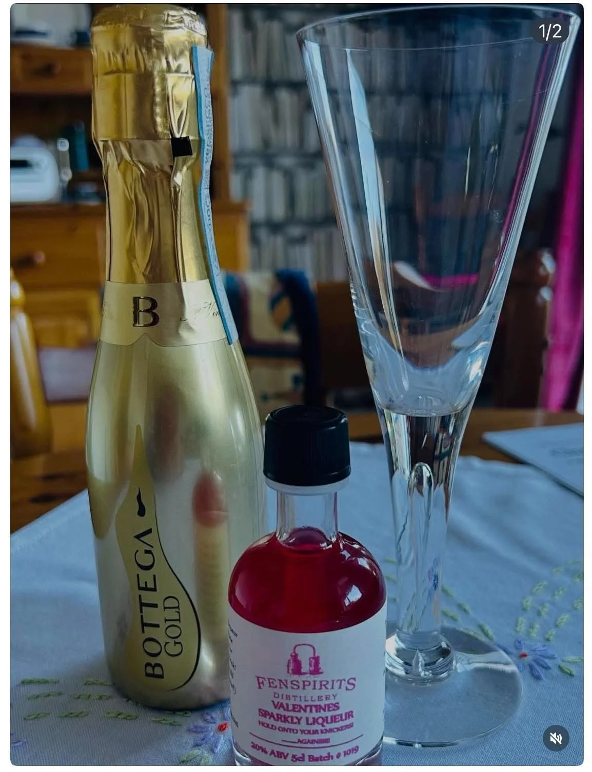 A bottle of Bottega Gold prosecco, a bottle of Fenspirit Valentines Sparkly Liquor, a tall empty champagne flute, and a small bottle of red liquid on a white embroidered tablecloth.