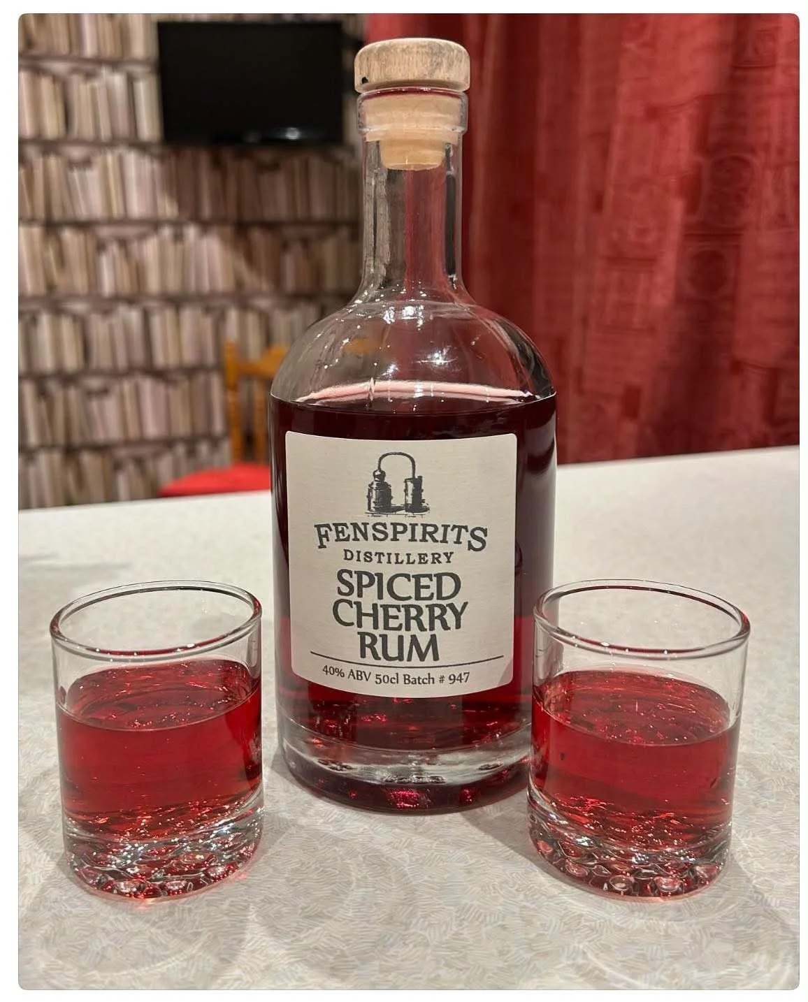 A bottle of Fenspirits Distillery spiced cherry rum with two glasses of red liquid on a white tablecloth, with a bookshelf and red curtain in the background.